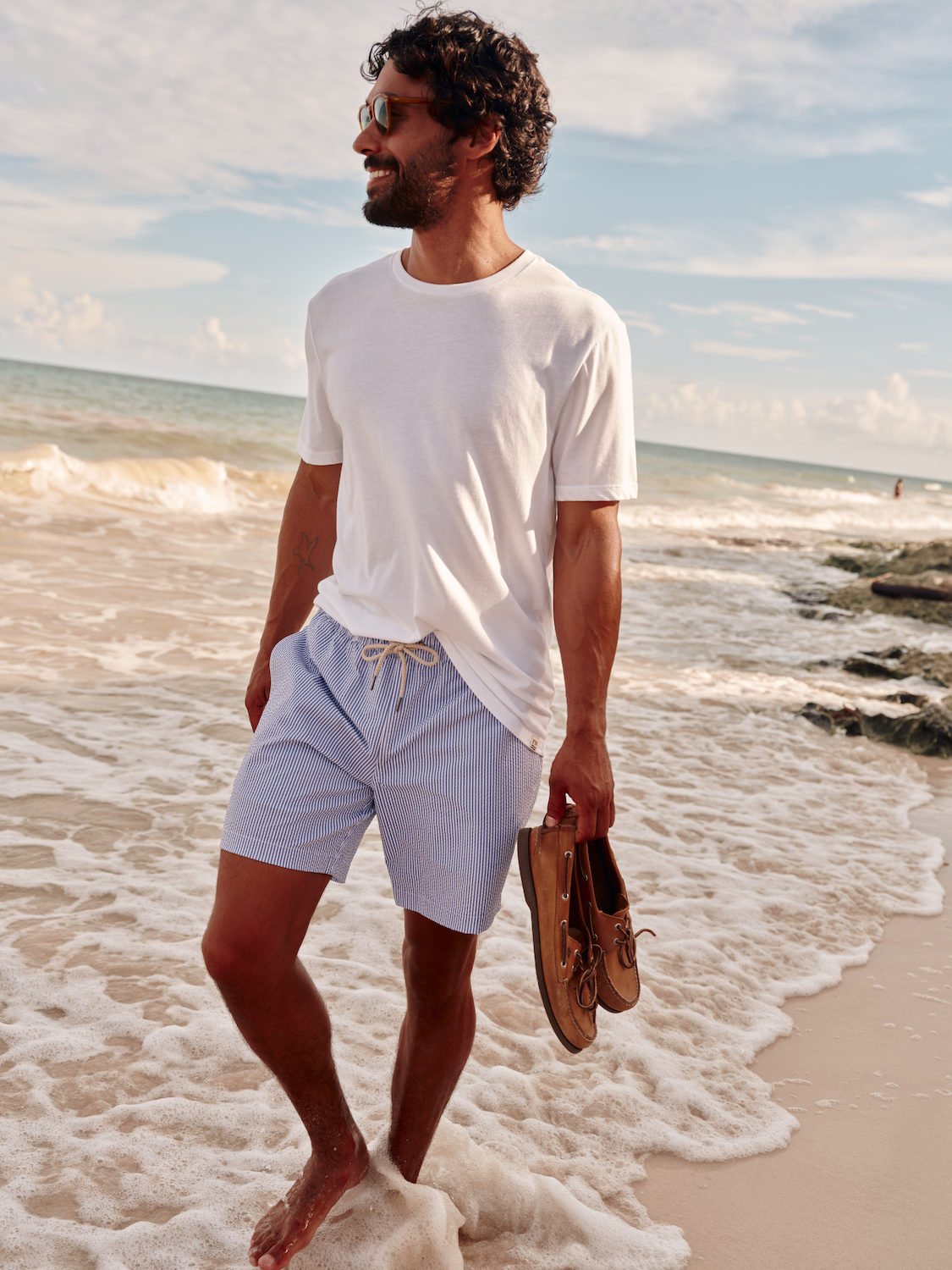 Man walking along water edge carrying his shoes while wearing the Bayberry Trunk and Saltaire tee