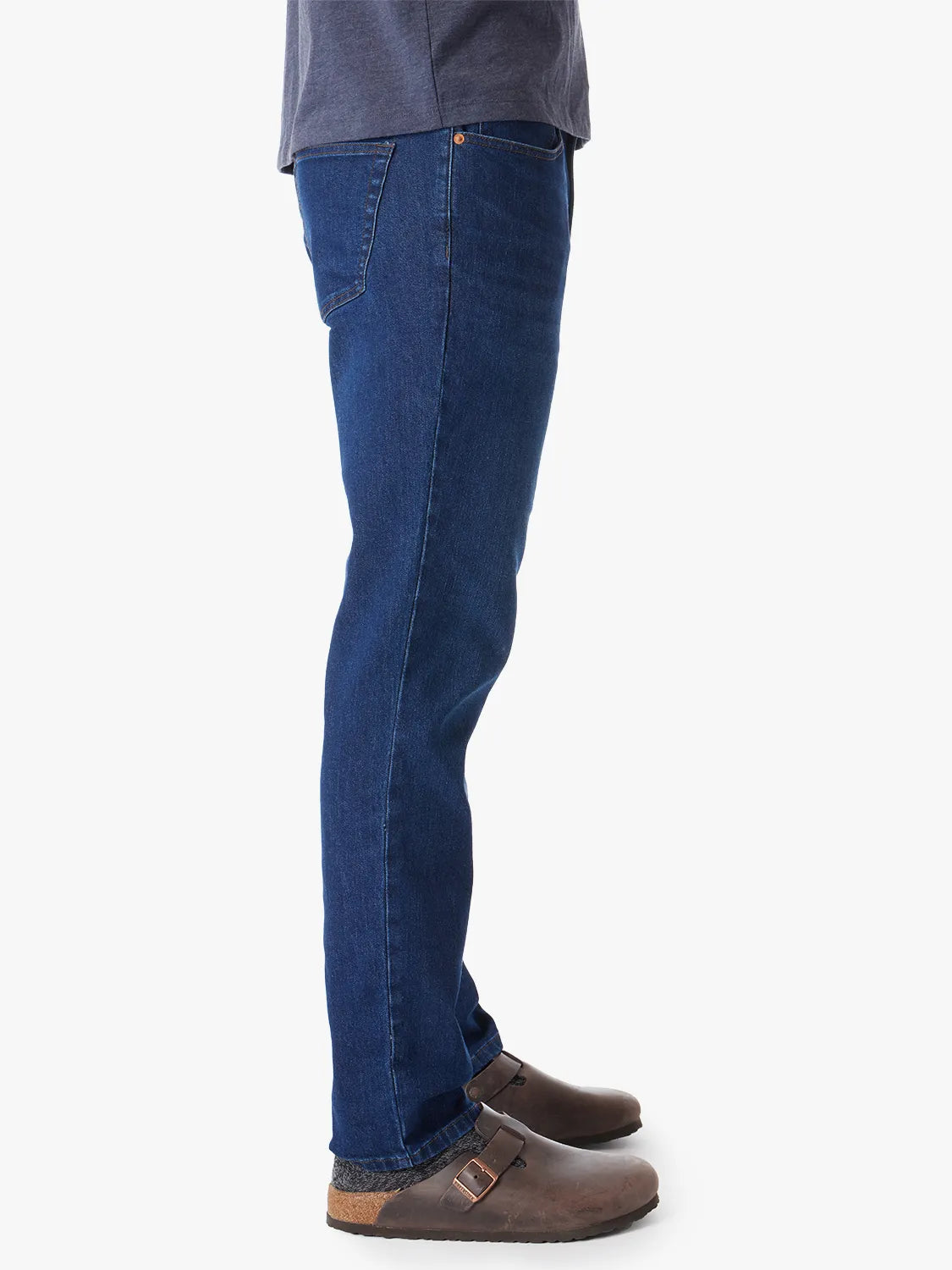 Person wearing a pair of men's straight-fit blue denim jeans in color Deep Sea Wash with a 5-pocket design on a white background. Paired with black tee and slide on shoes, showing the side view.