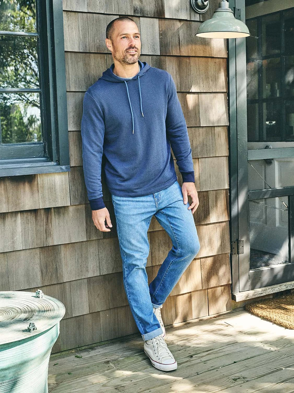Man wearing driftwood denim jeans in beach wash color paired with navy tailwind hoodie and sneakers while standing outside house.