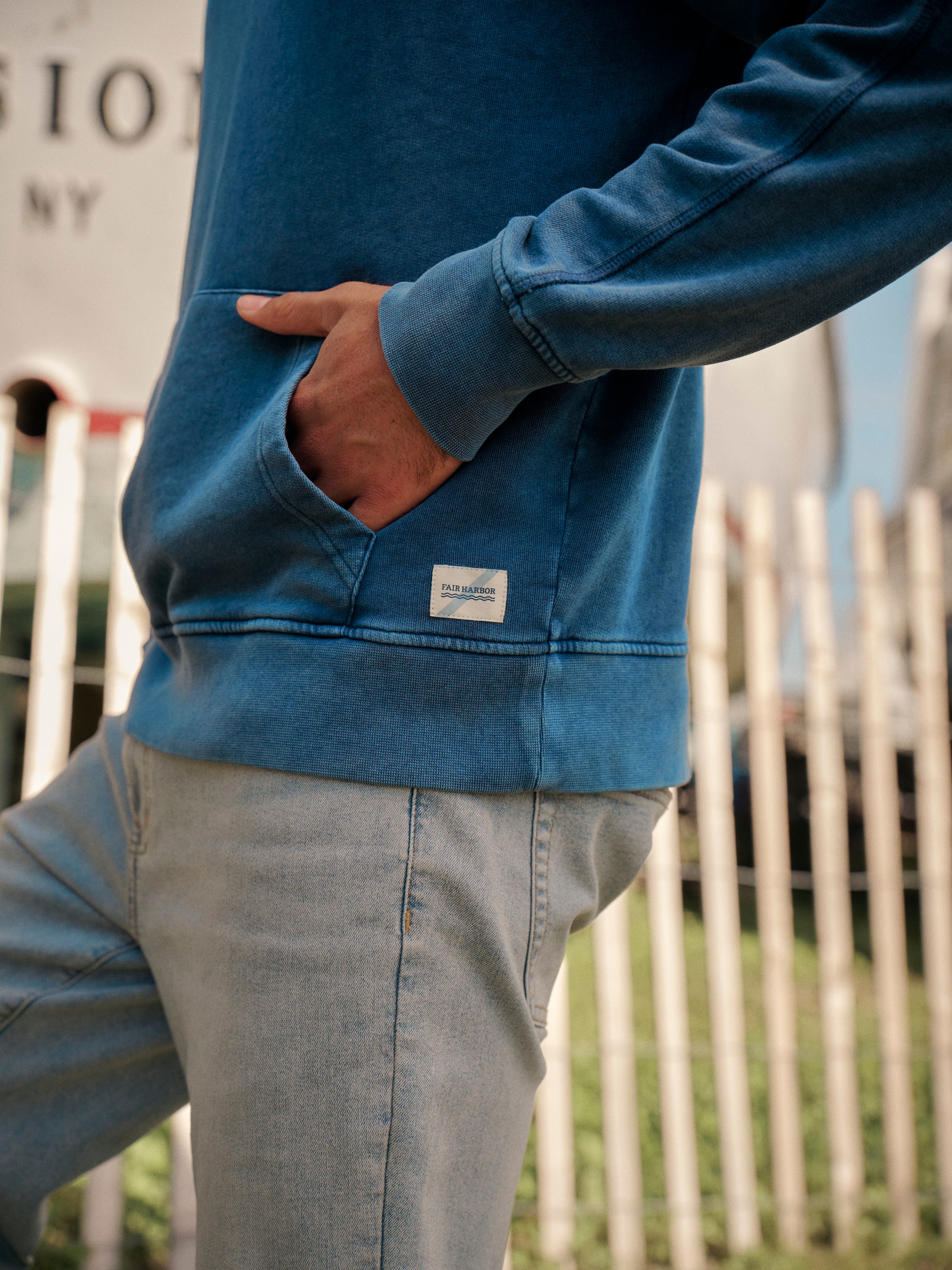 Person wearing a blue saltaire hoodie with a hand in kangaroo pocket and a visible brand logo, standing outdoors by fence.