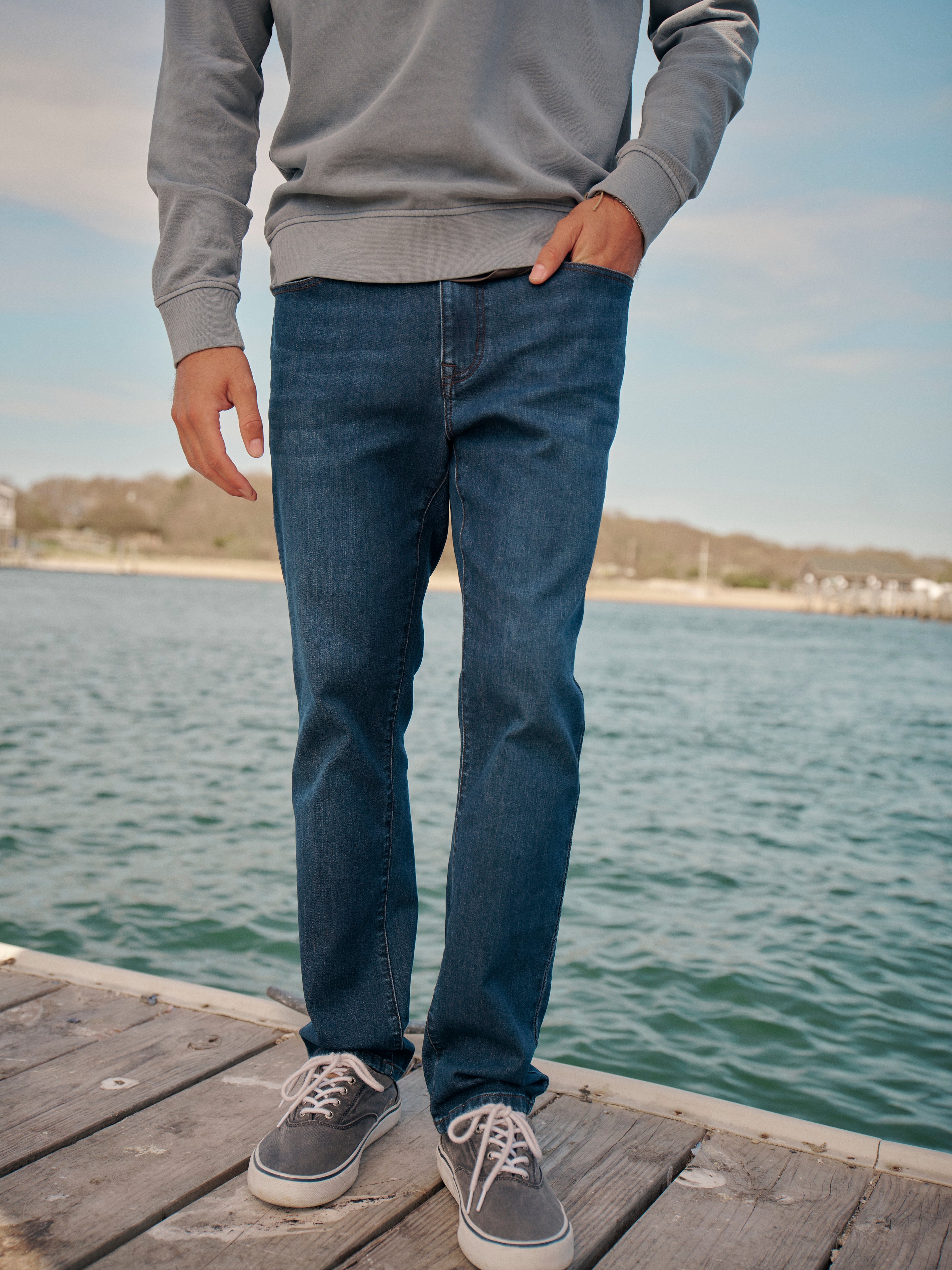 Person wearing deep sea wash colored jeans and a gray saltaire crewneck standing on a wooden dock by a body of water.