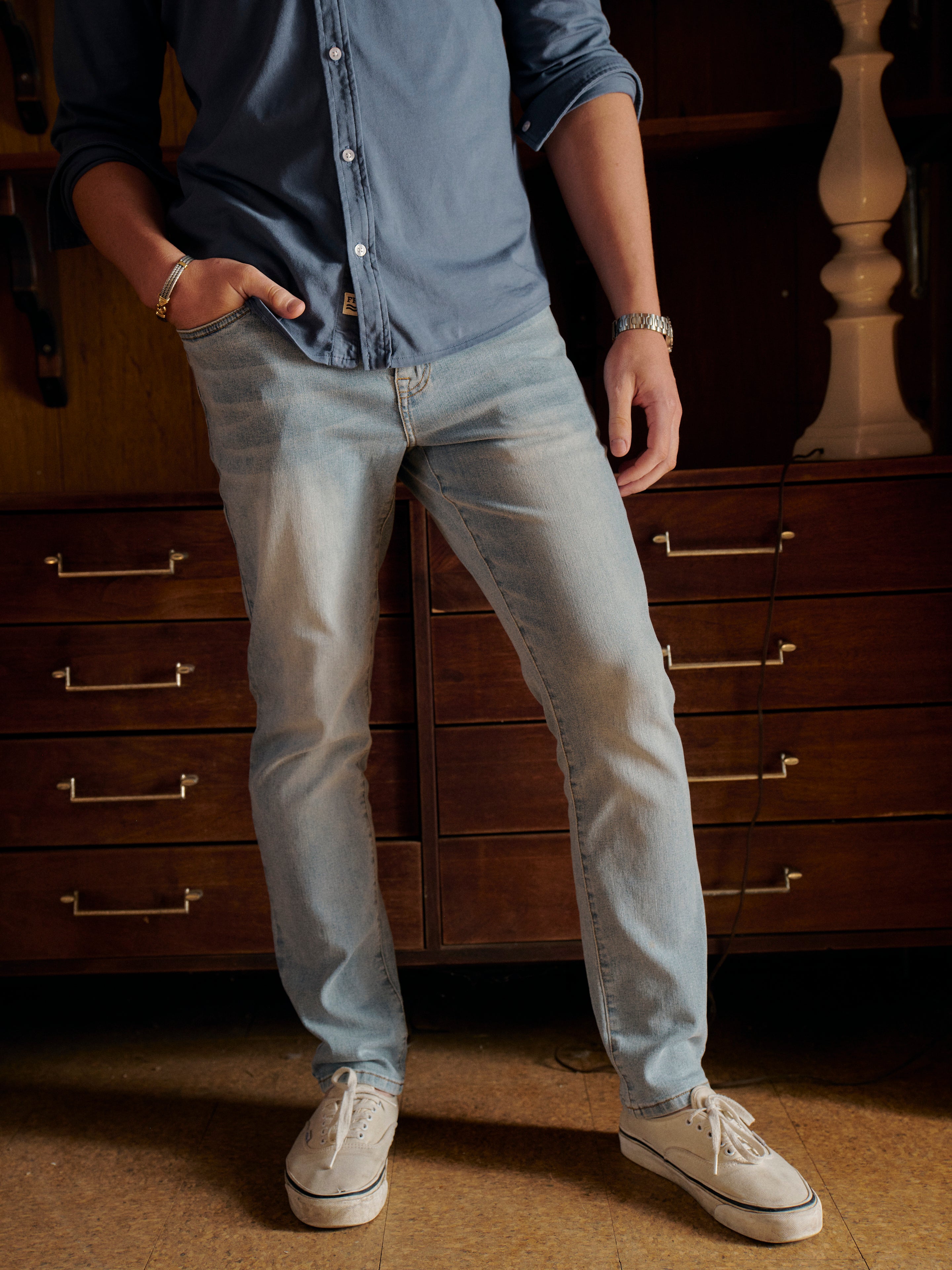 Person wearing a blue saltaire long sleeve shirt and light wash driftwood jeans standing in front of wooden drawers.