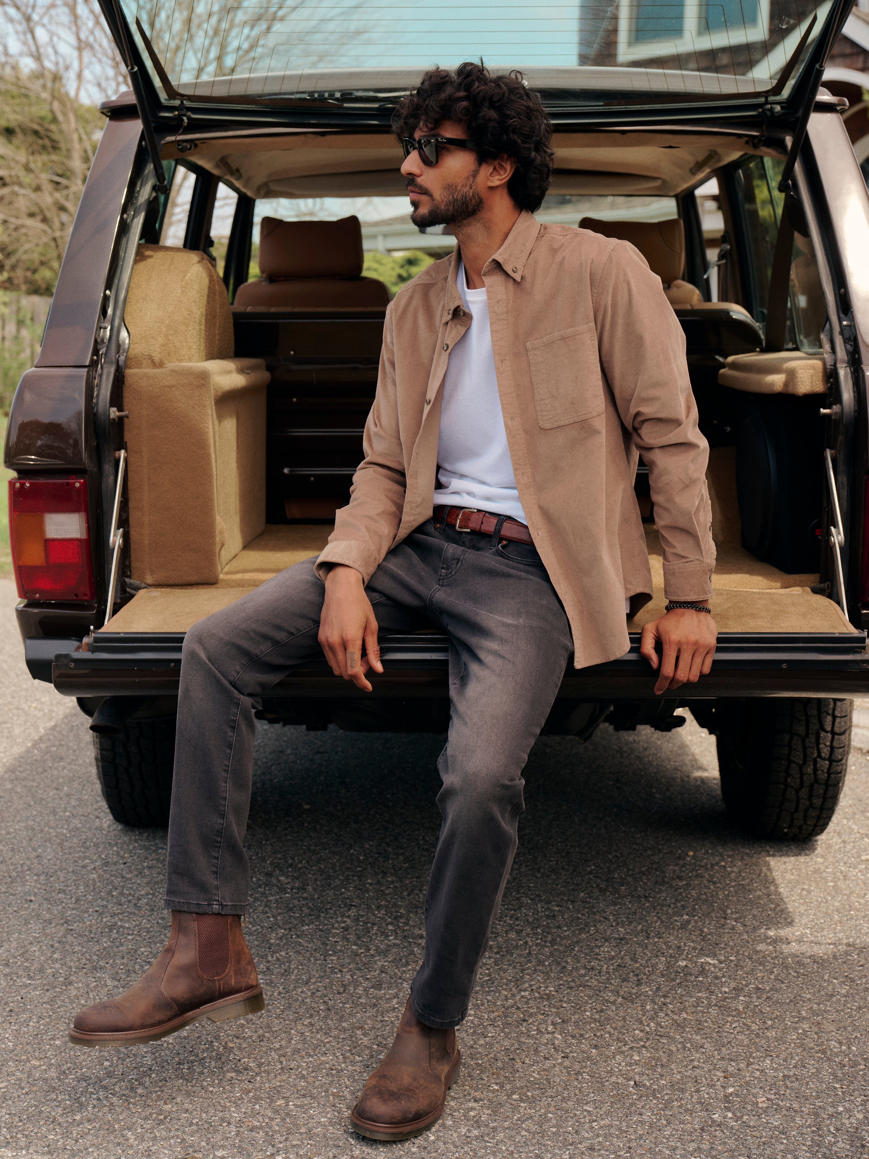 Man sitting in the open trunk of a car wearing a Wilder Stretch Corduroy Shirt, white Saltaire Tshirt and black Driftwood denim pants.