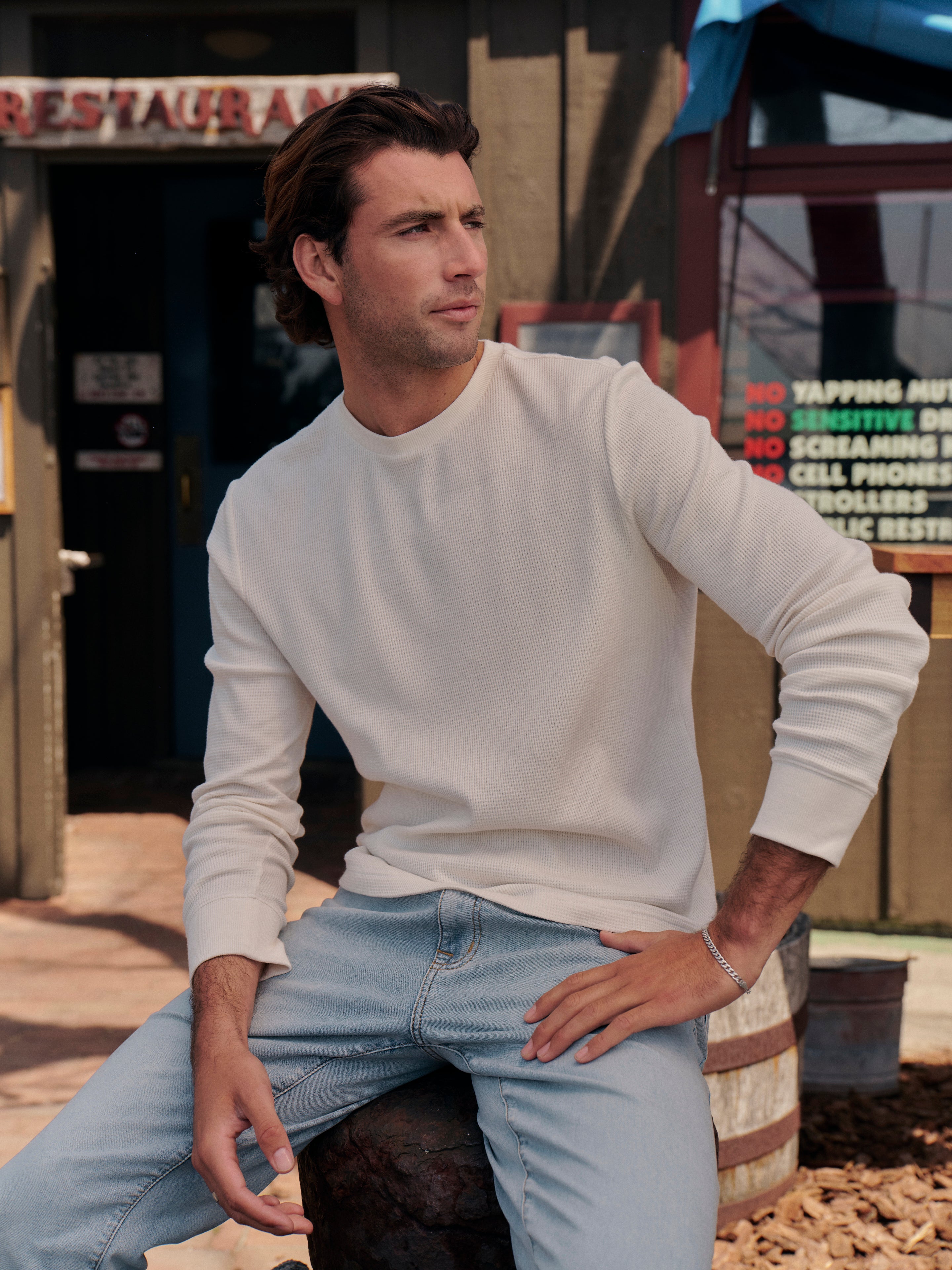 Man wearing a cream waffle long sleeve shirt and blue jeans sitting on a bench with a restaurant sign in the background.