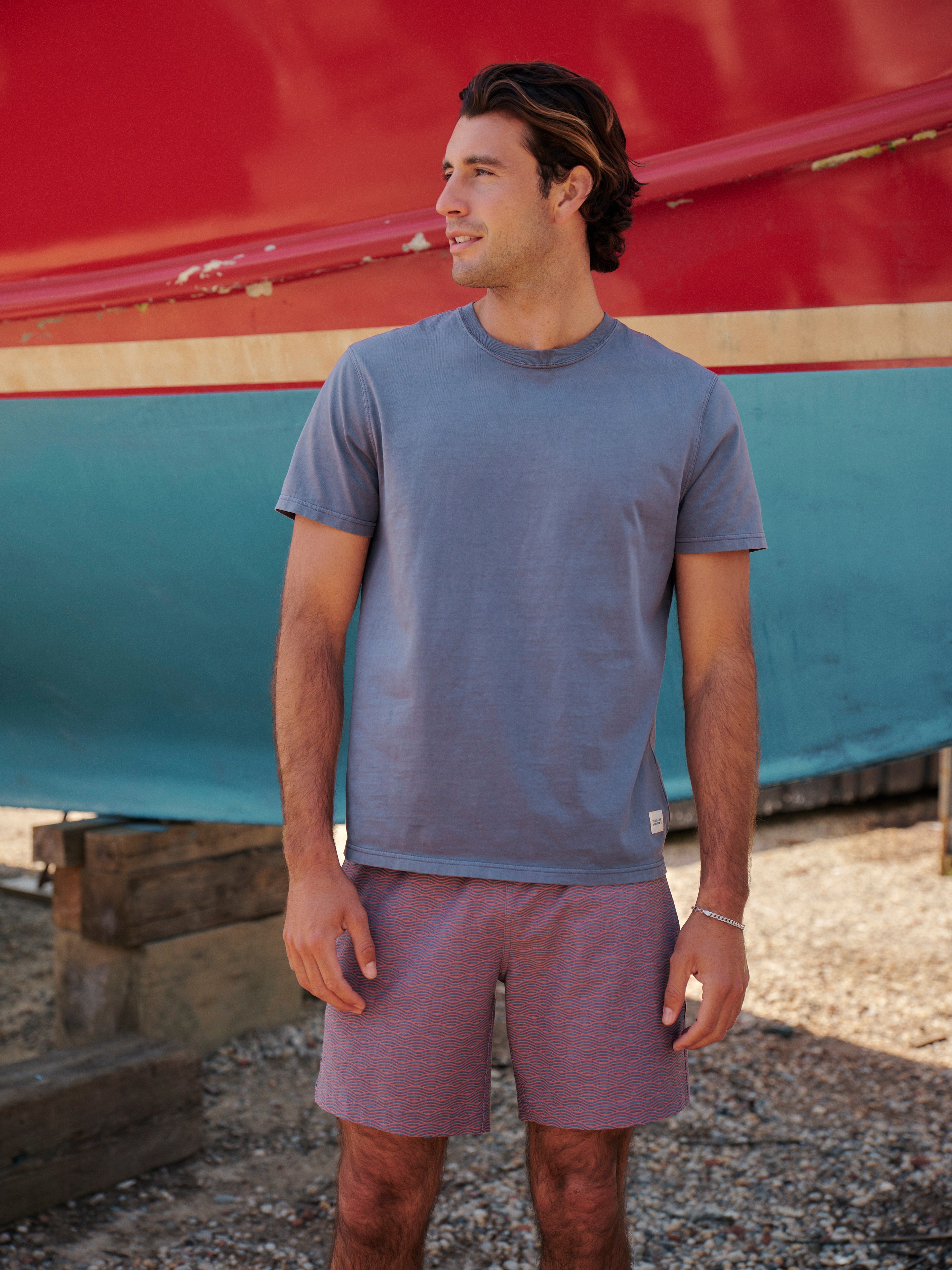 Man wearing a blue saltaire t-shirt and bayberry trunks standing in front of a colorful boat.