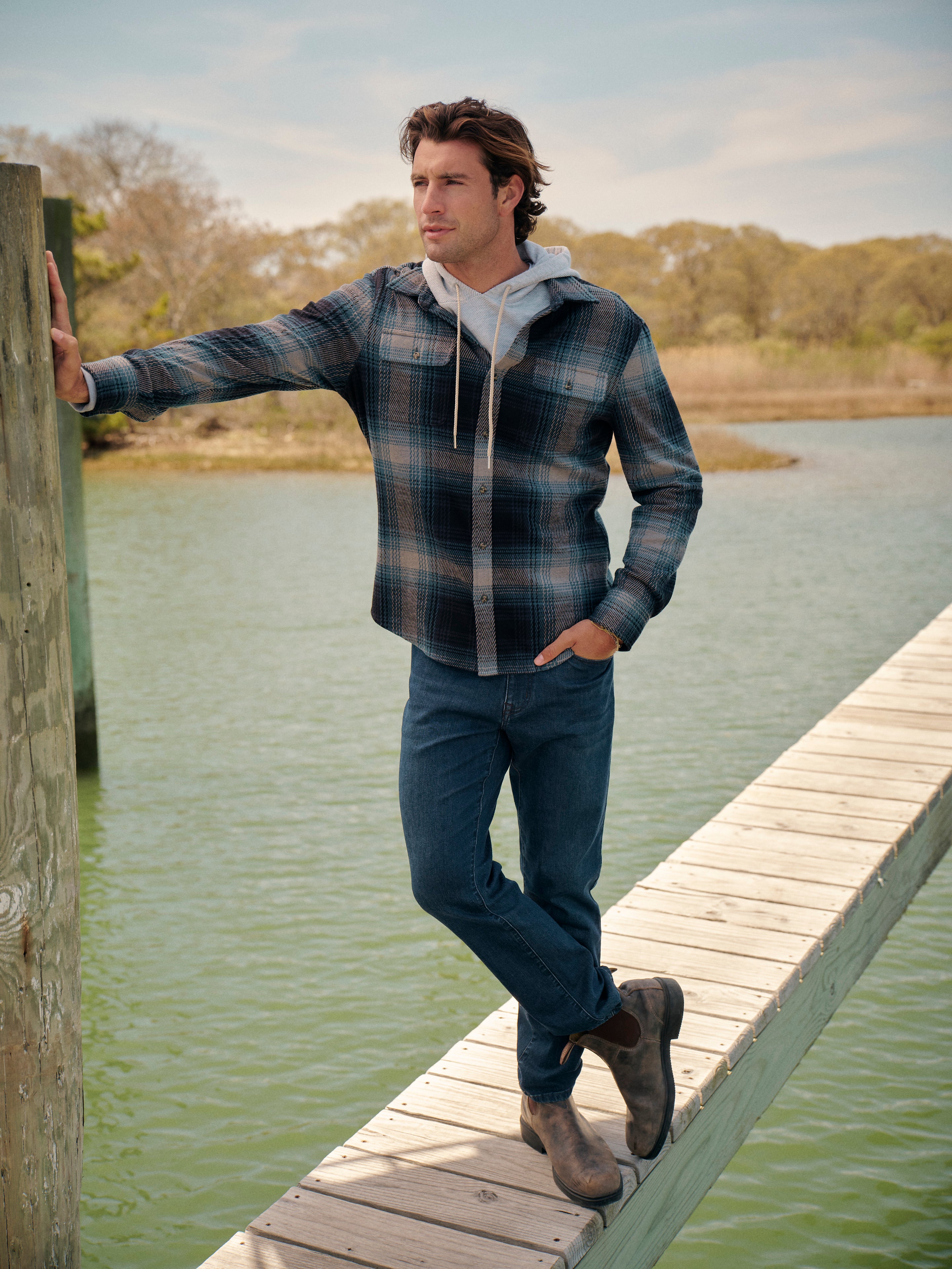 Man standing on a wooden dock by a body of water wearing a Dunewood flannel layered with tailwood hoodie  and deep sea wash colored jeans.