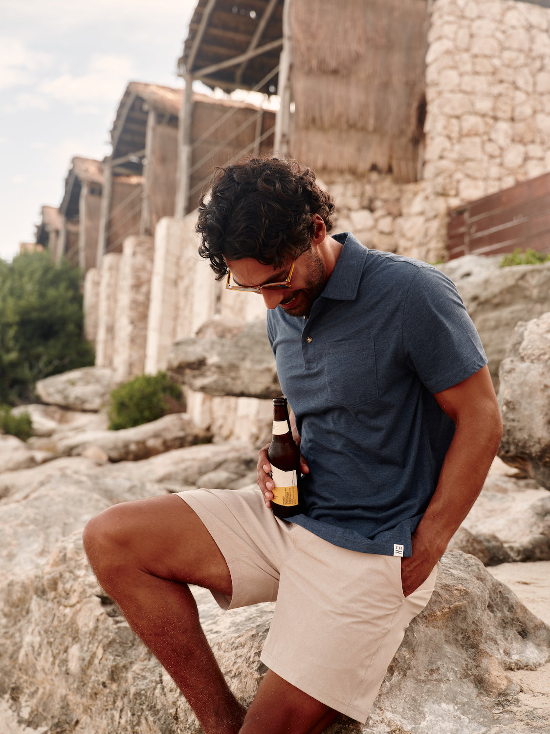 Man leaning on boulder wearing the khkai one short and a navy polo with drink in his hand along coastline.
