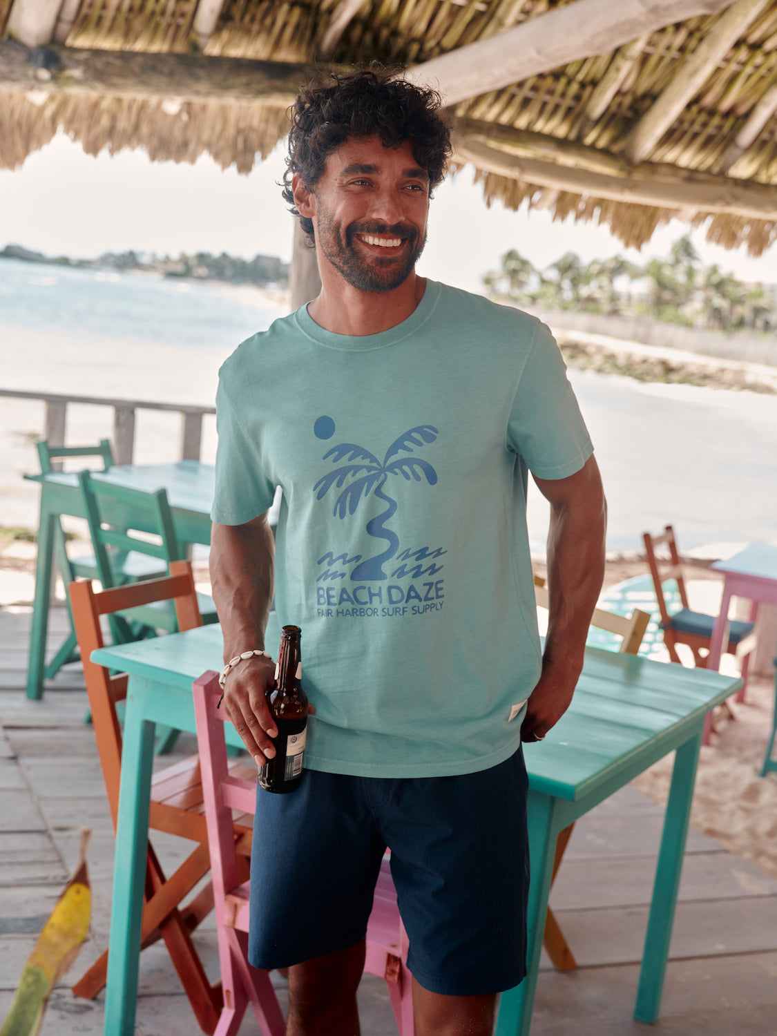 Man standing on patio wearing navy one short and tshirt, with drink in hand. Colorful table and chairs behind him.