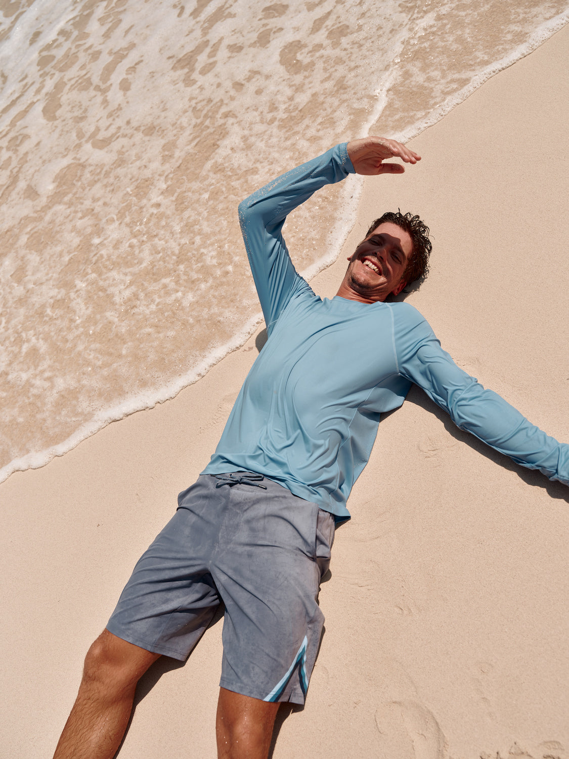 Man laying down in the sand, along the edge of water on the beach. Wearing a ozone board trunk and anchor rashguard.