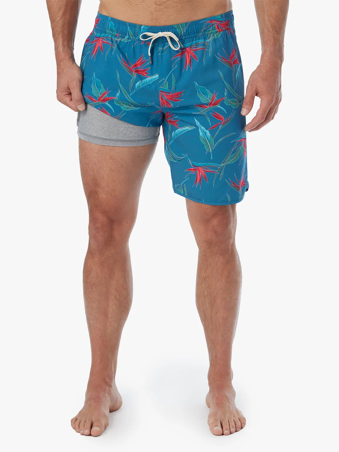A person wearing a pair of teal blue Anchor Trunk with a red and green bird of paradise print with a drawstring waist, showing the liner on a white background. 