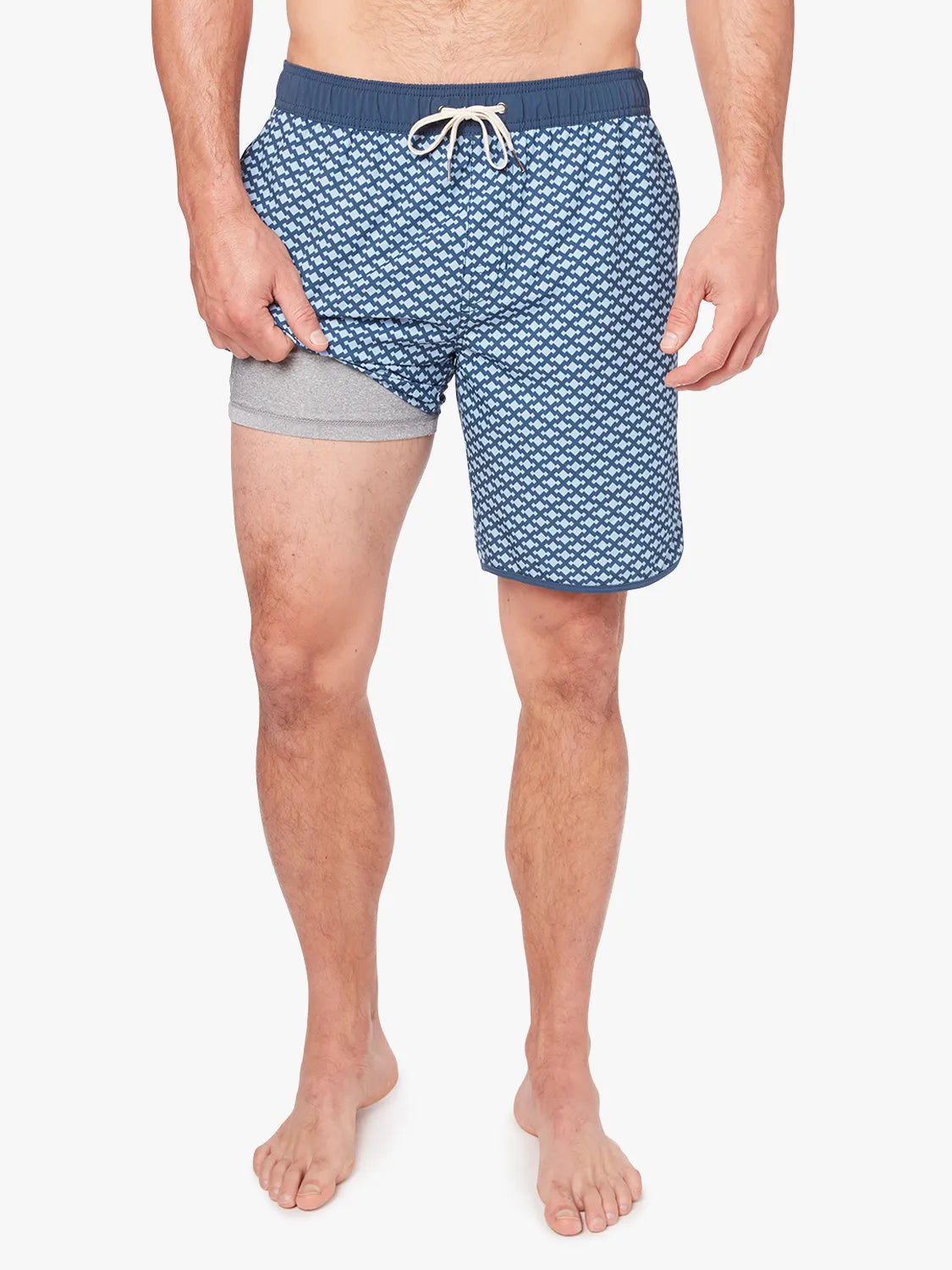 A person wearing a pair of Anchor Trunk in 8 inch inseam with a geometric pattern in shades of blue, featuring a drawstring waist showing the liner against a white background. 