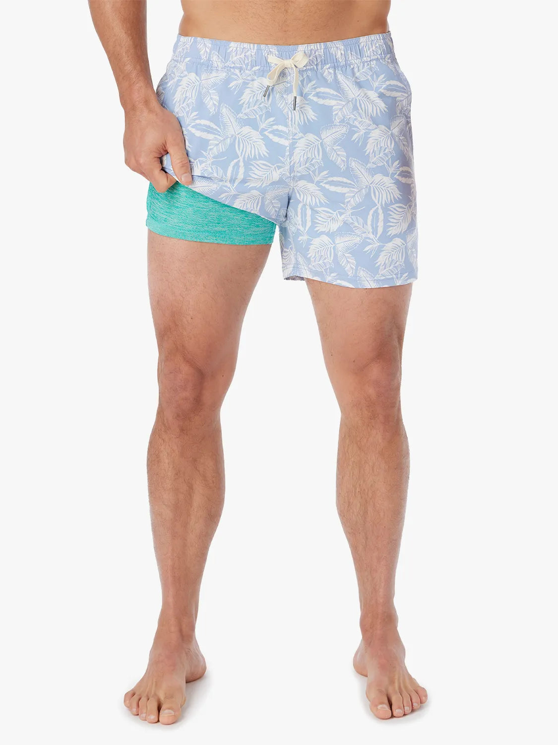 Person wearing light blue bungalow swim trunks with white tropical leaf pattern on a white background showing the liner. 