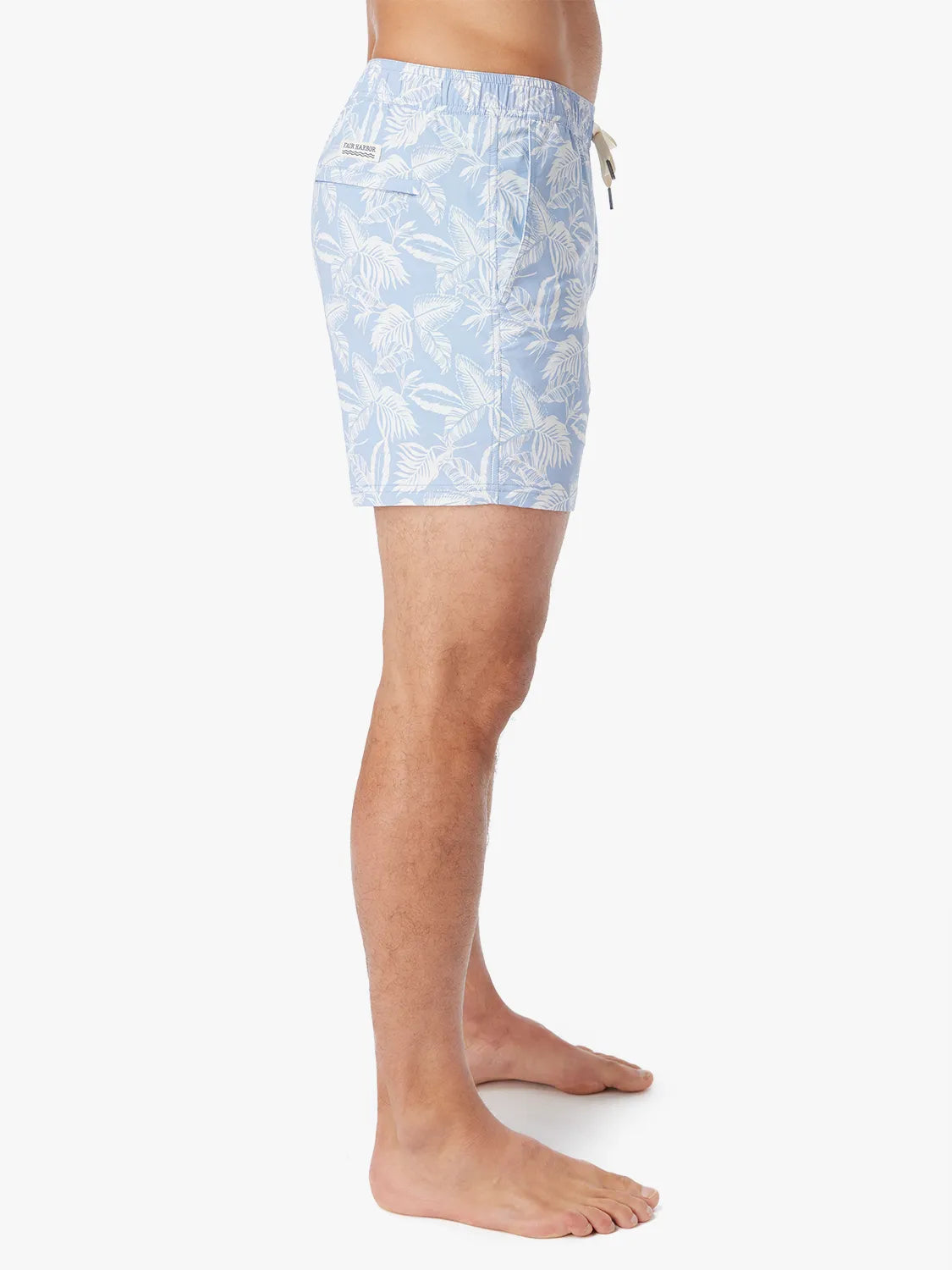 Person wearing light blue bungalow swim trunks with white tropical leaf pattern on a white background showing the side view. 