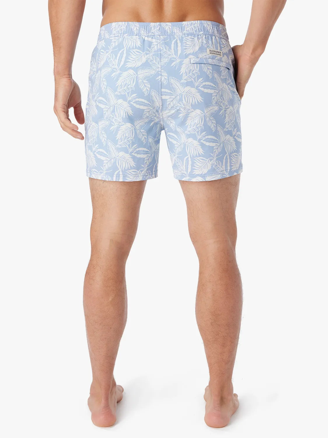 Person wearing light blue bungalow swim trunks with white tropical leaf pattern on a white background showing the back view. 