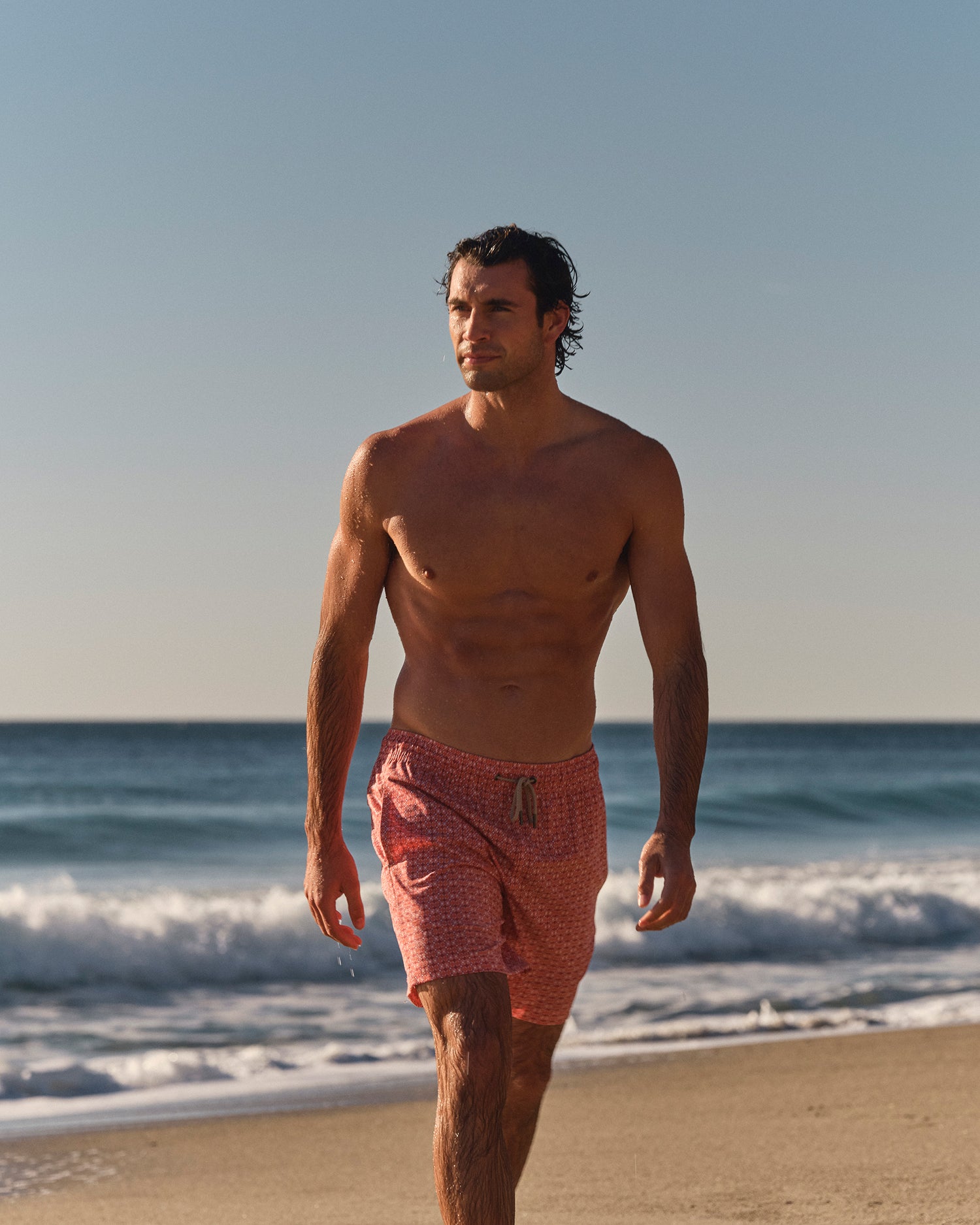 Man walking on a beach wearing red and orange swim shorts with a pattern.