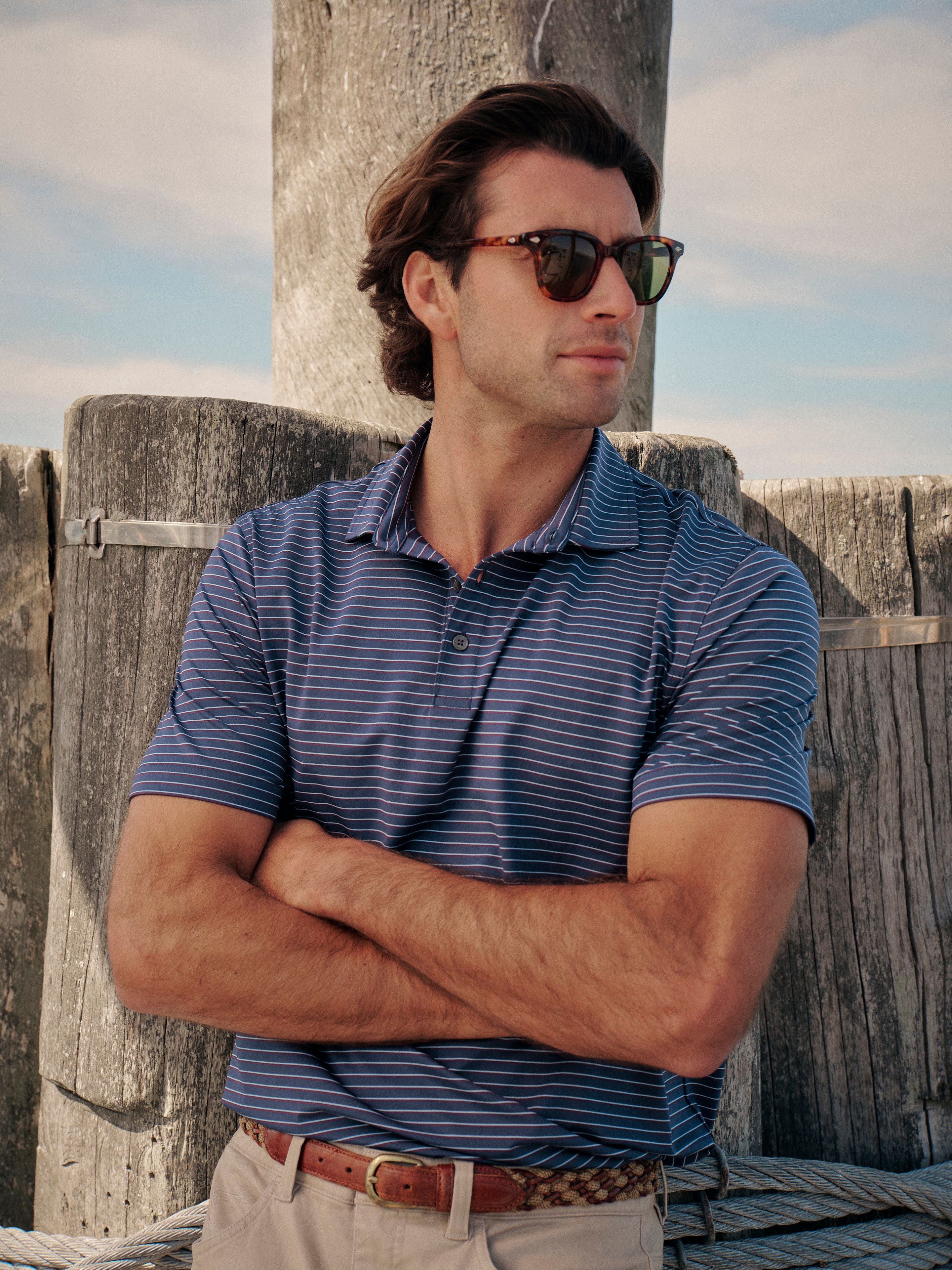 Man wearing sunglasses and a striped compass polo shirt standing against a wooden fence.