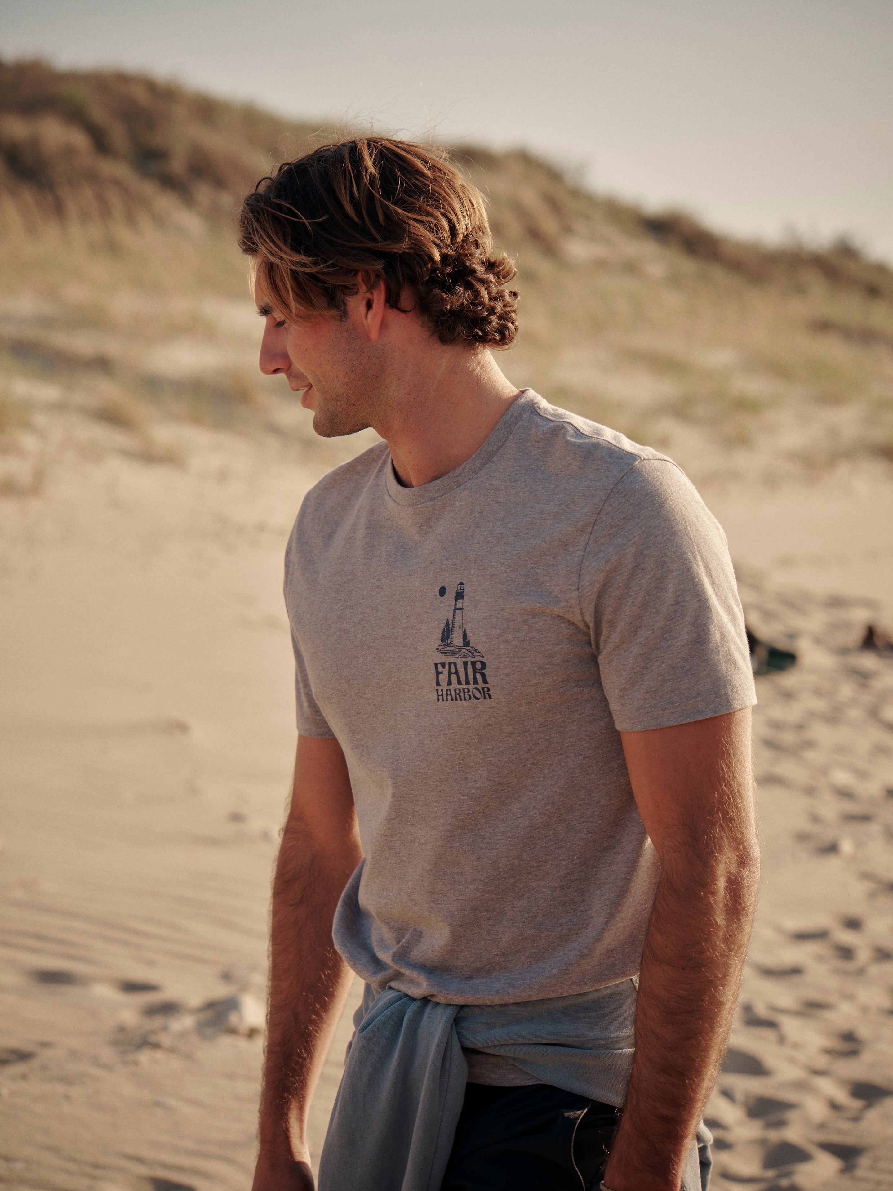 Man wearing a grey kismet t-shirt with a logo on right chest on a sandy beach