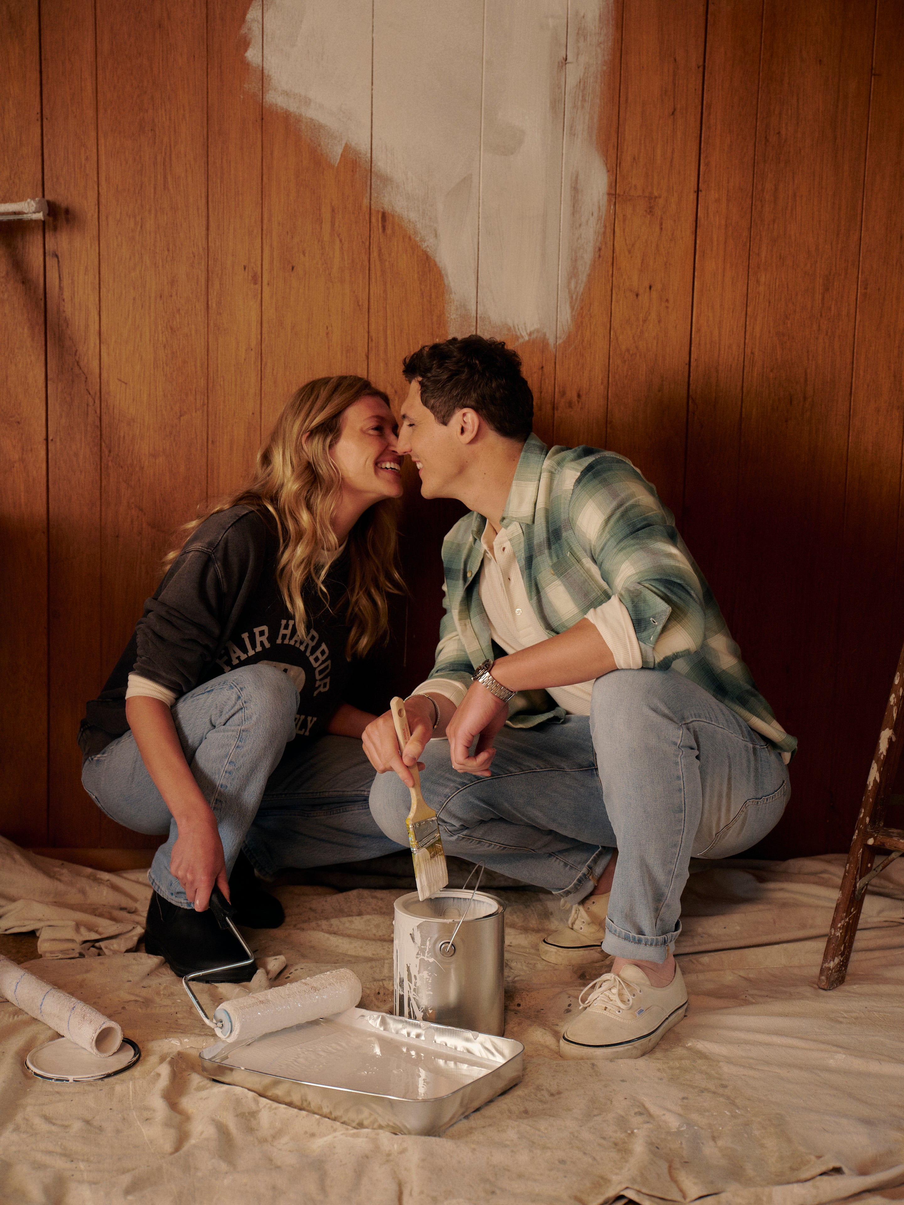 Two people sitting on the floor with painting supplies against a wooden wall.