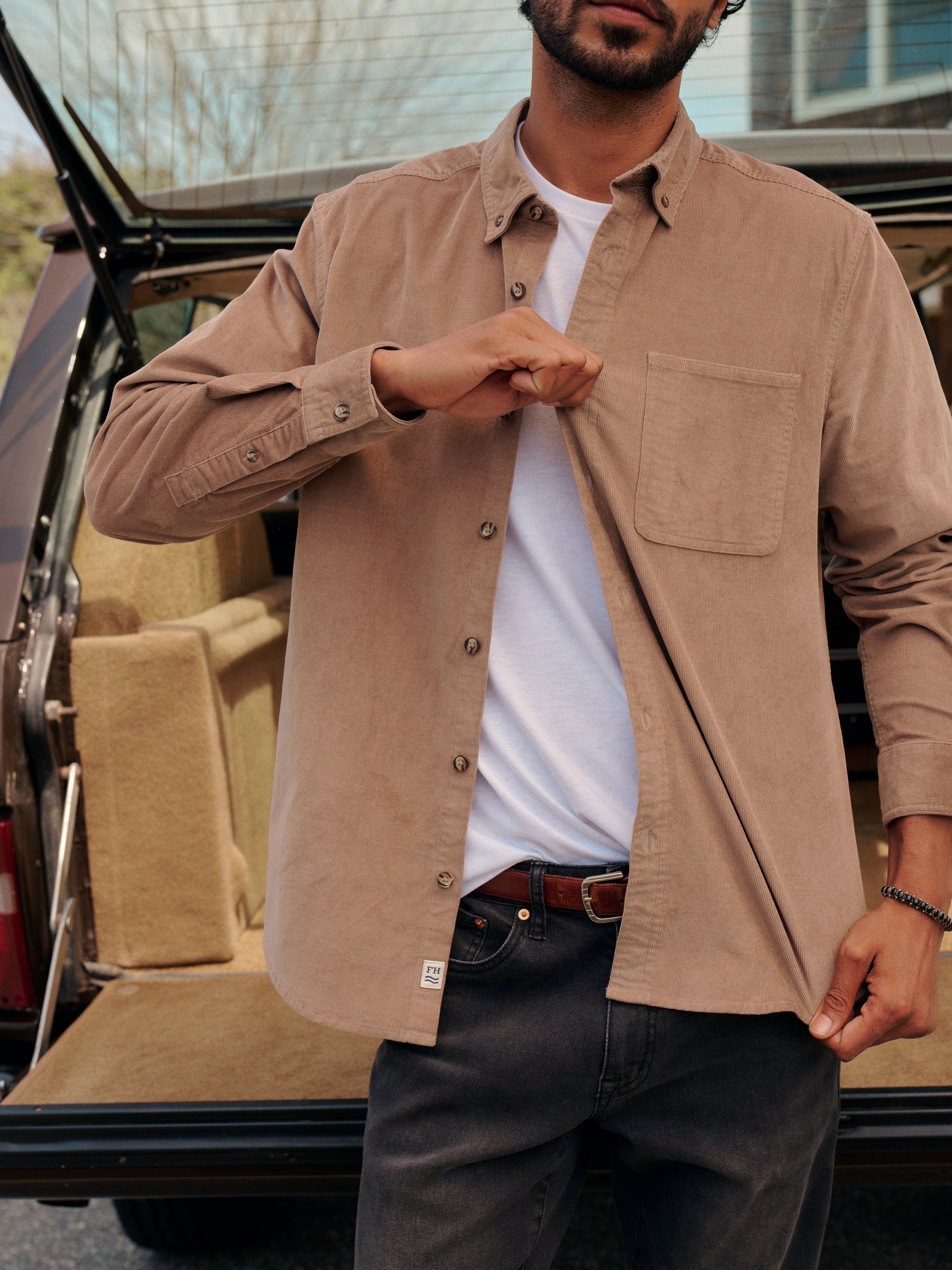 Man standing in front of an open trunk of car with a house in the background. Wearing brown wilder corduroy shirt and black driftwood denim. Man is pulling on the shirt fabric to demonstrate the fabrics stretching ability. 