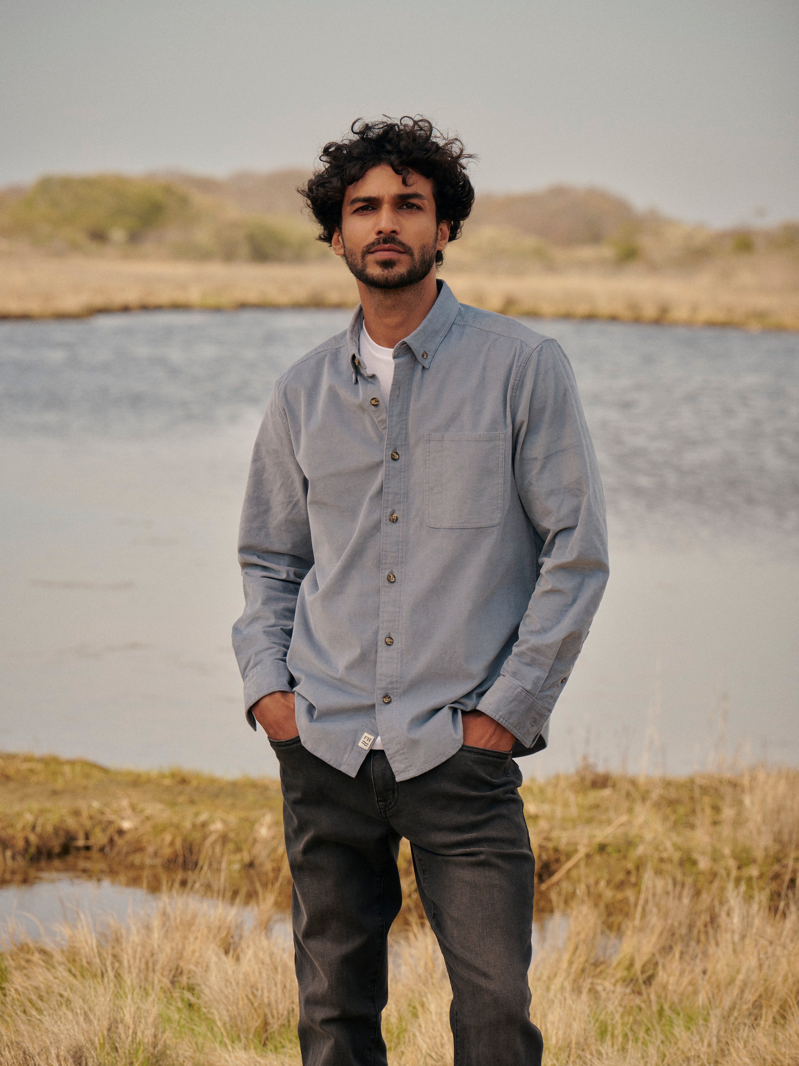 Man standing by a body of water with grassy areas, wearing wilder corduroy shirt in maritime and driftwood denim in black. 