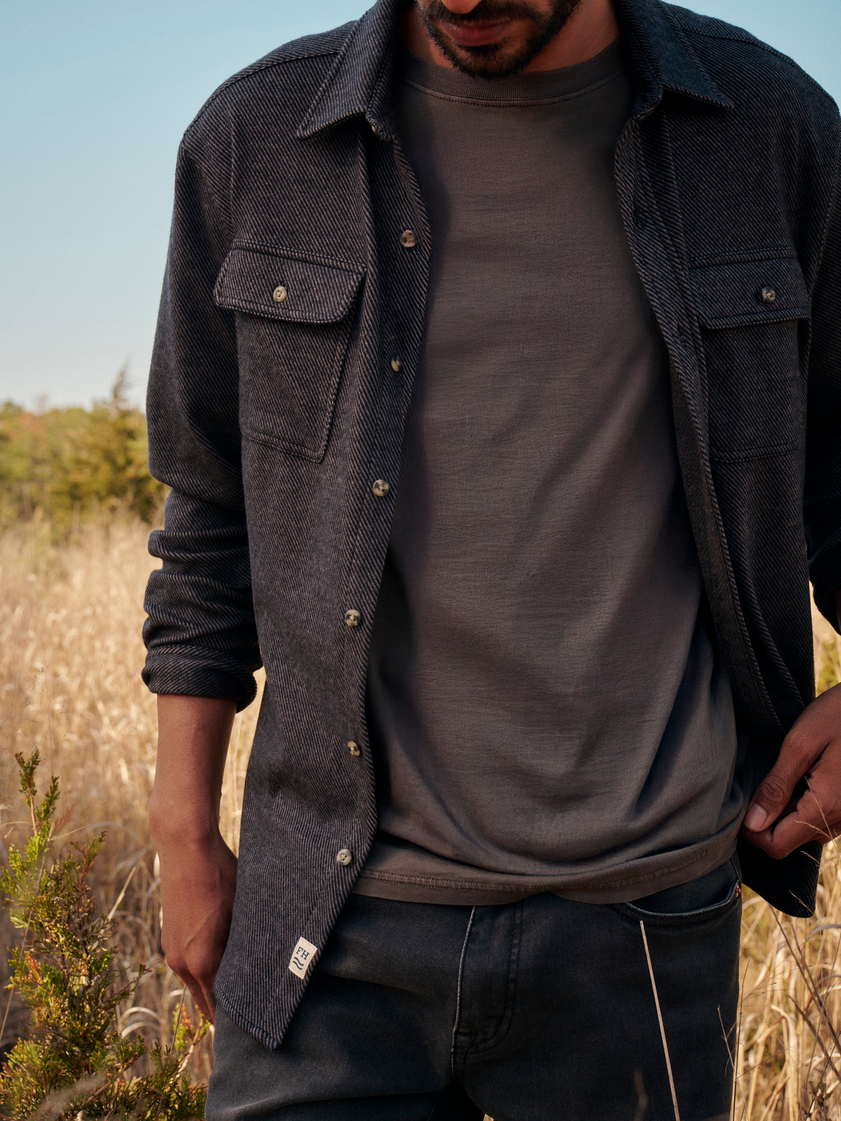 Man wearing a black dunewood twill flannel  over a black saltaire t-shirt in a natural setting with trees and clear sky.