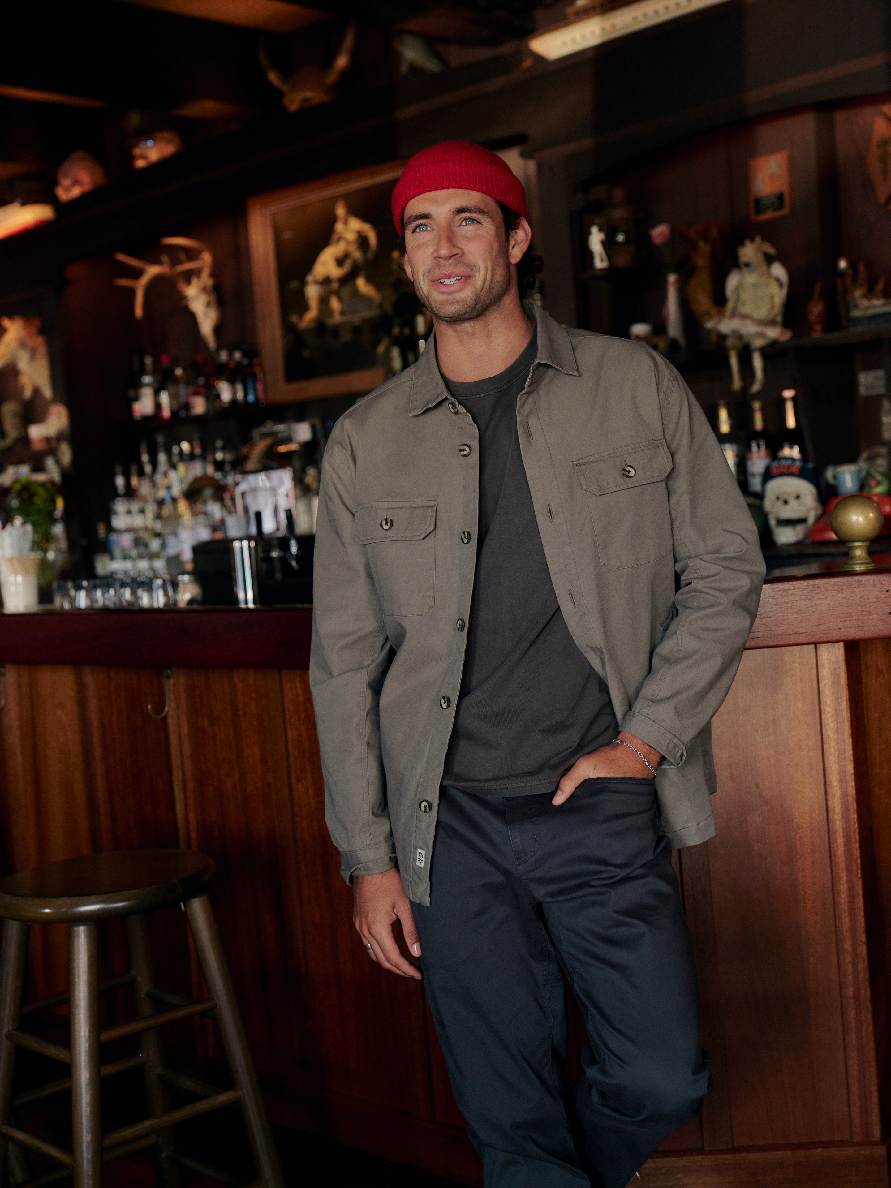 Man standing in a bar with wooden decor and shelves in the background wearing olive green montauk jacket with a red beanie and black pants. 