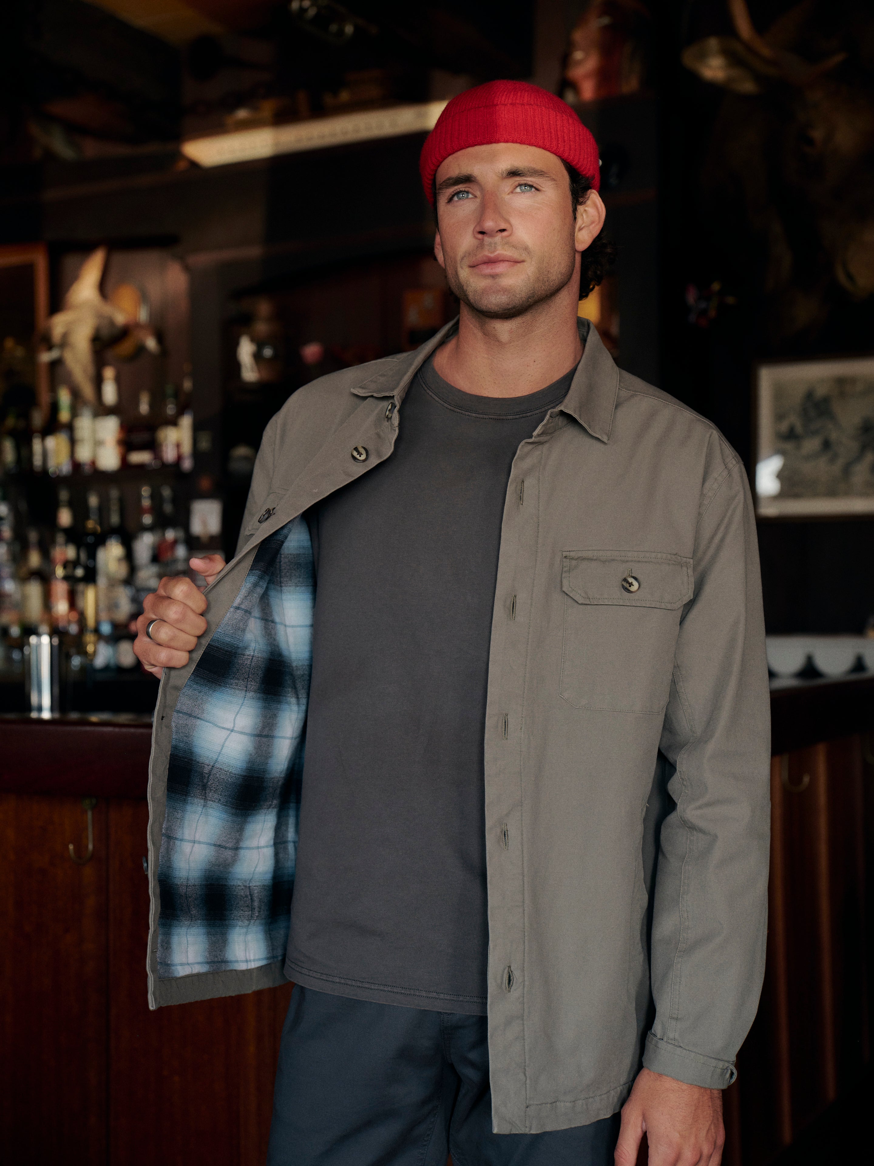 Man standing in a bar with wooden decor and shelves in the background wearing olive green montauk jacket with a red beanie and black pants. Showing the plaid lining of olive jacket. 