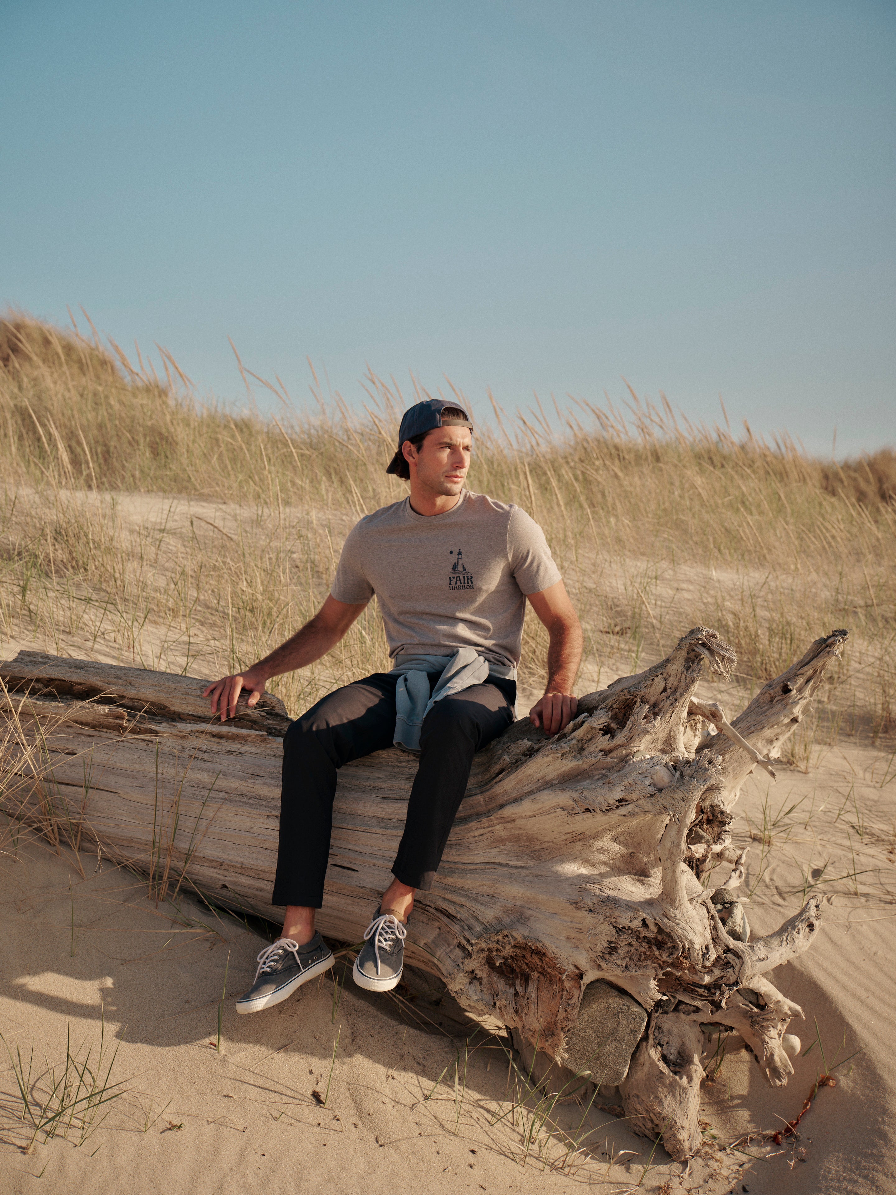 Man sitting on a log in a sandy dune landscape wearing kismet t-shirt and black pants with a backwards baseball  hat. 