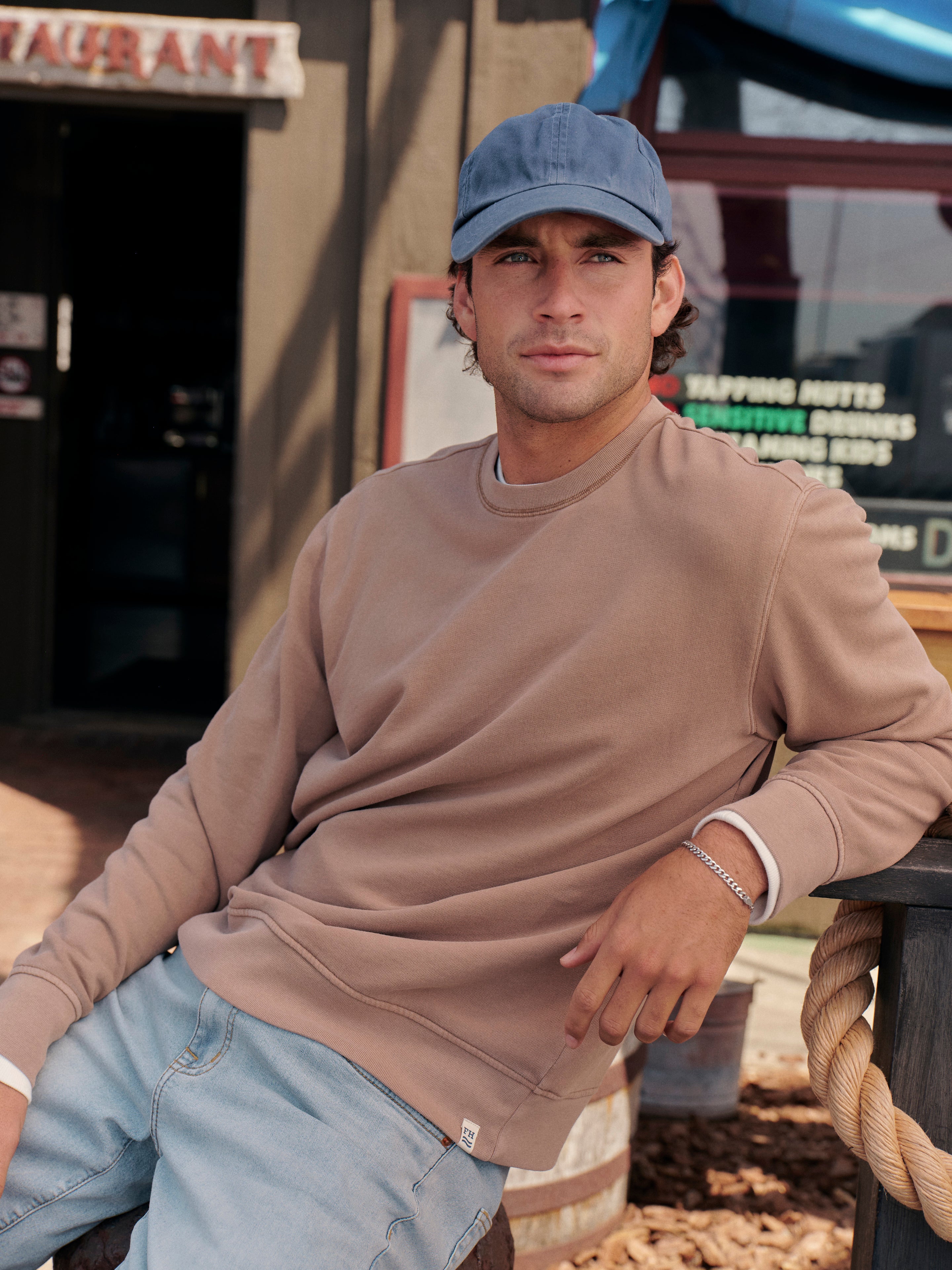 Man sitting in front of restaurant with visible signs in the background. Wearing a baseball hat, saltaire sweatshirt and pair of jeans. 