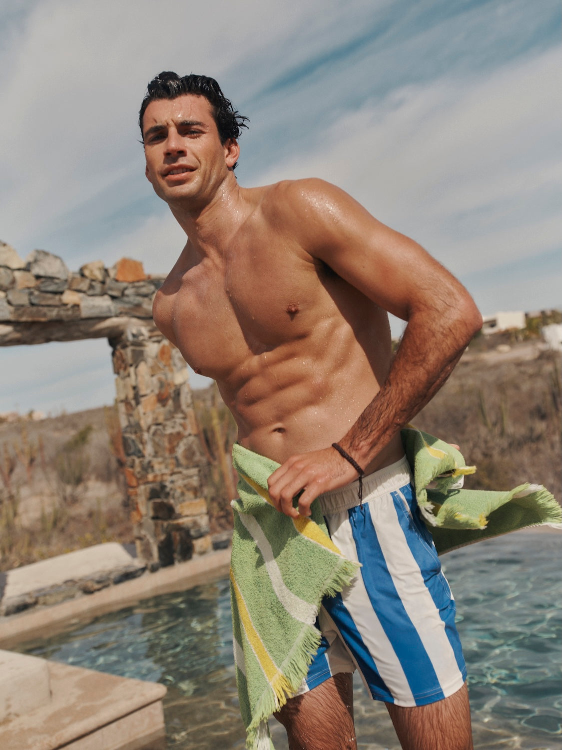 Man standing beside pool while drying off with a green towel, wearing the bungalow trunk.