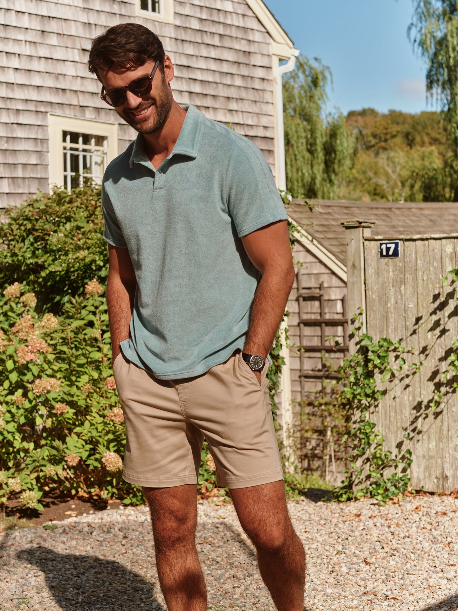 Man wearing a teal polo shirt and beige shorts standing outdoors near a wooden building.