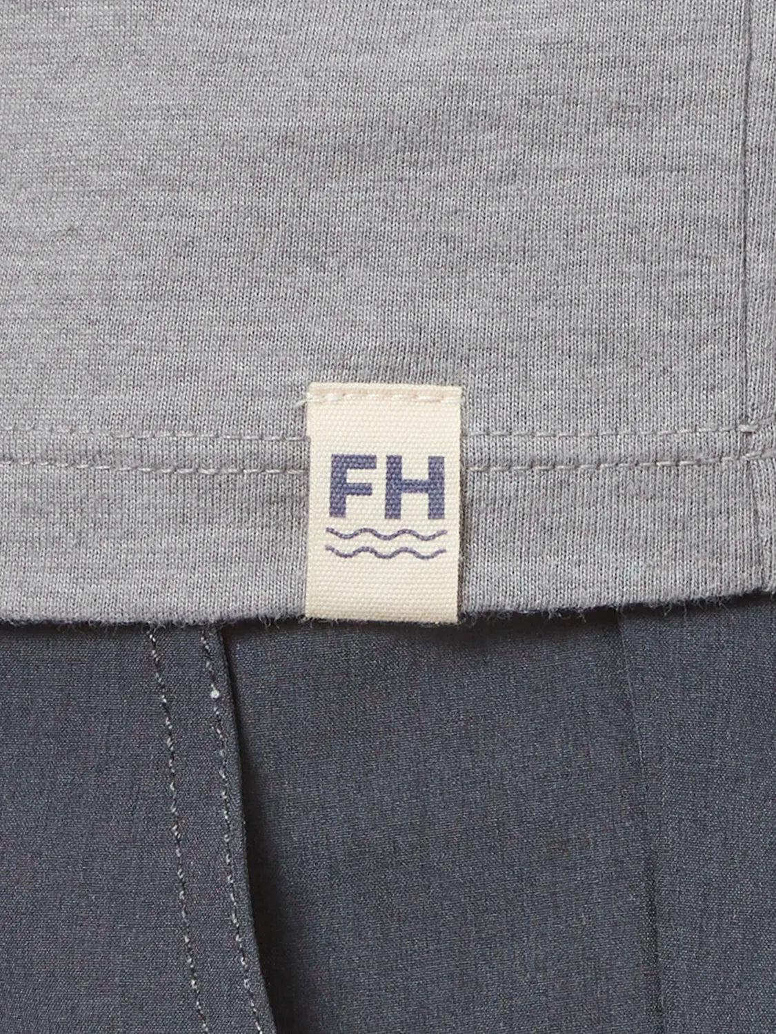 Close up of grey kismet t-shirt fabric and brand logo patch on bottom hem, paired with black one pants. 