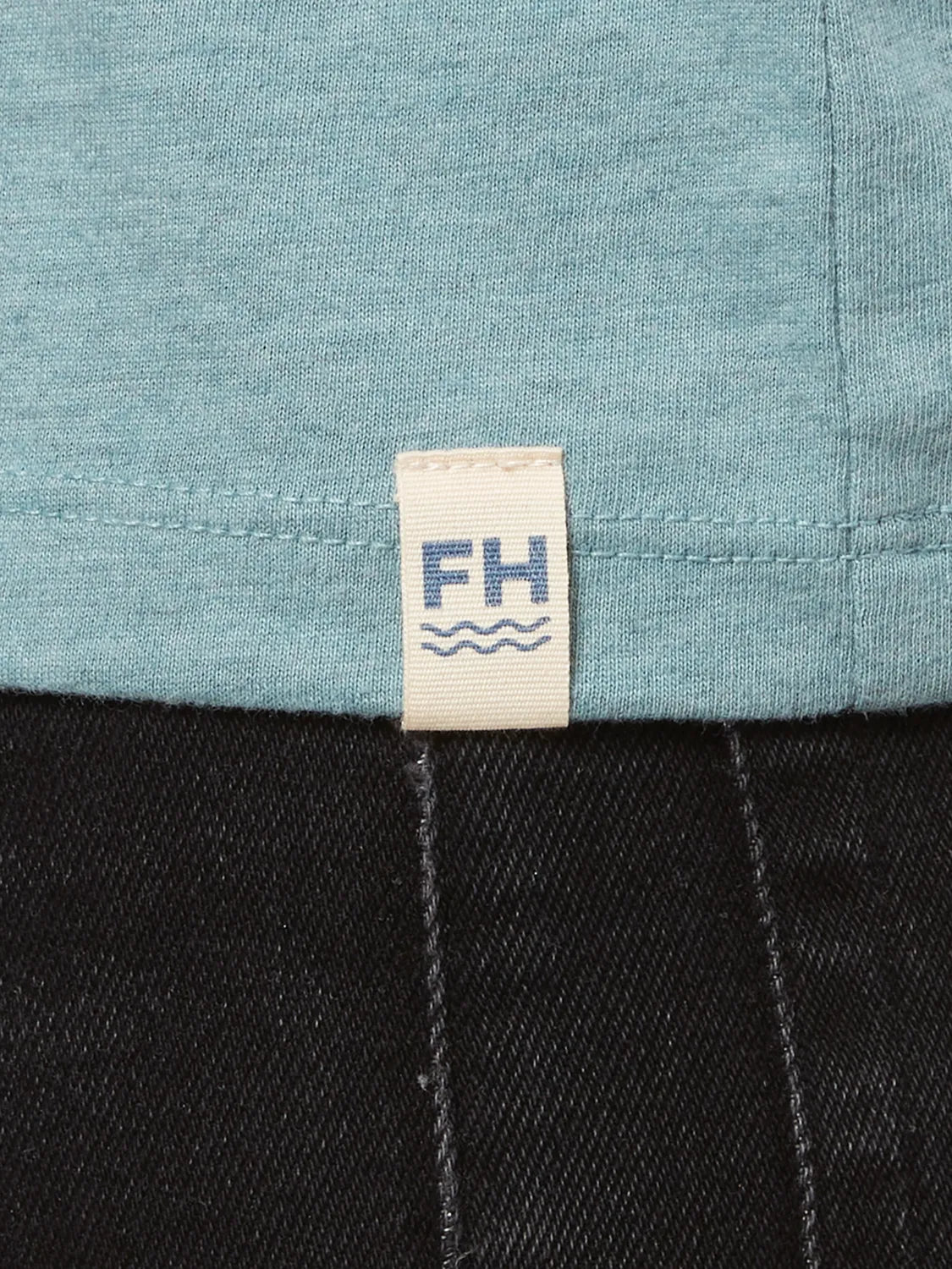 Close up of heather light green kismet t-shirt fabric and brand logo patch at bottom hem.