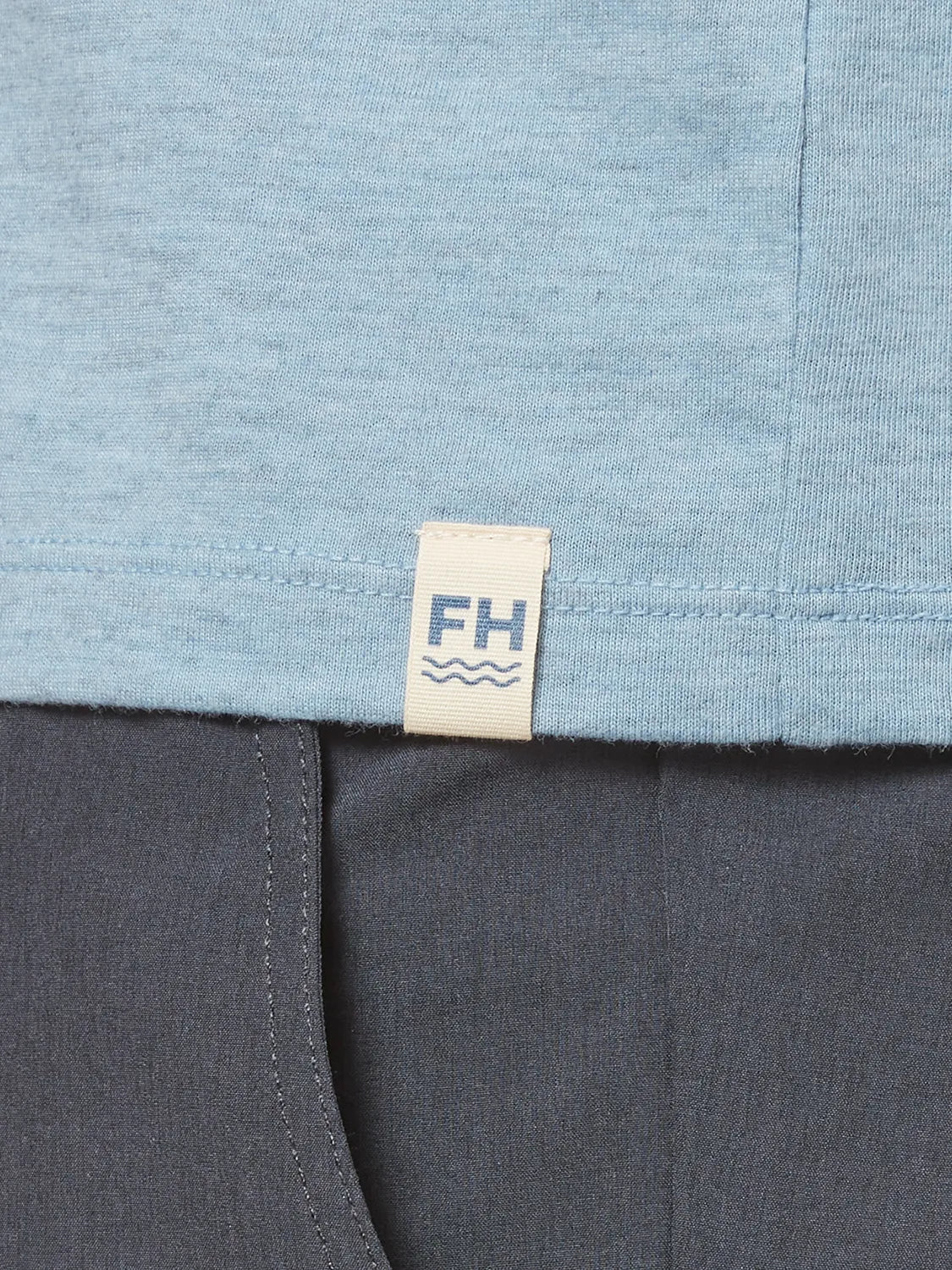 Close up of light blue kismet tee fabric and brand logo patch on bottom hem with black pants. 