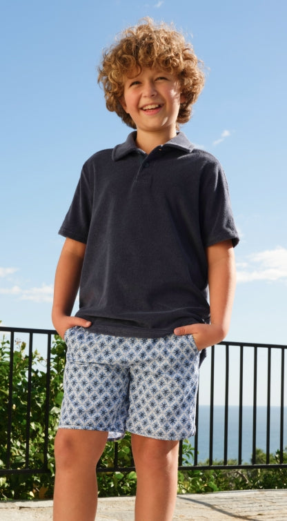Child wearing a navy polo shirt and patterned shorts standing outdoors with a clear sky.