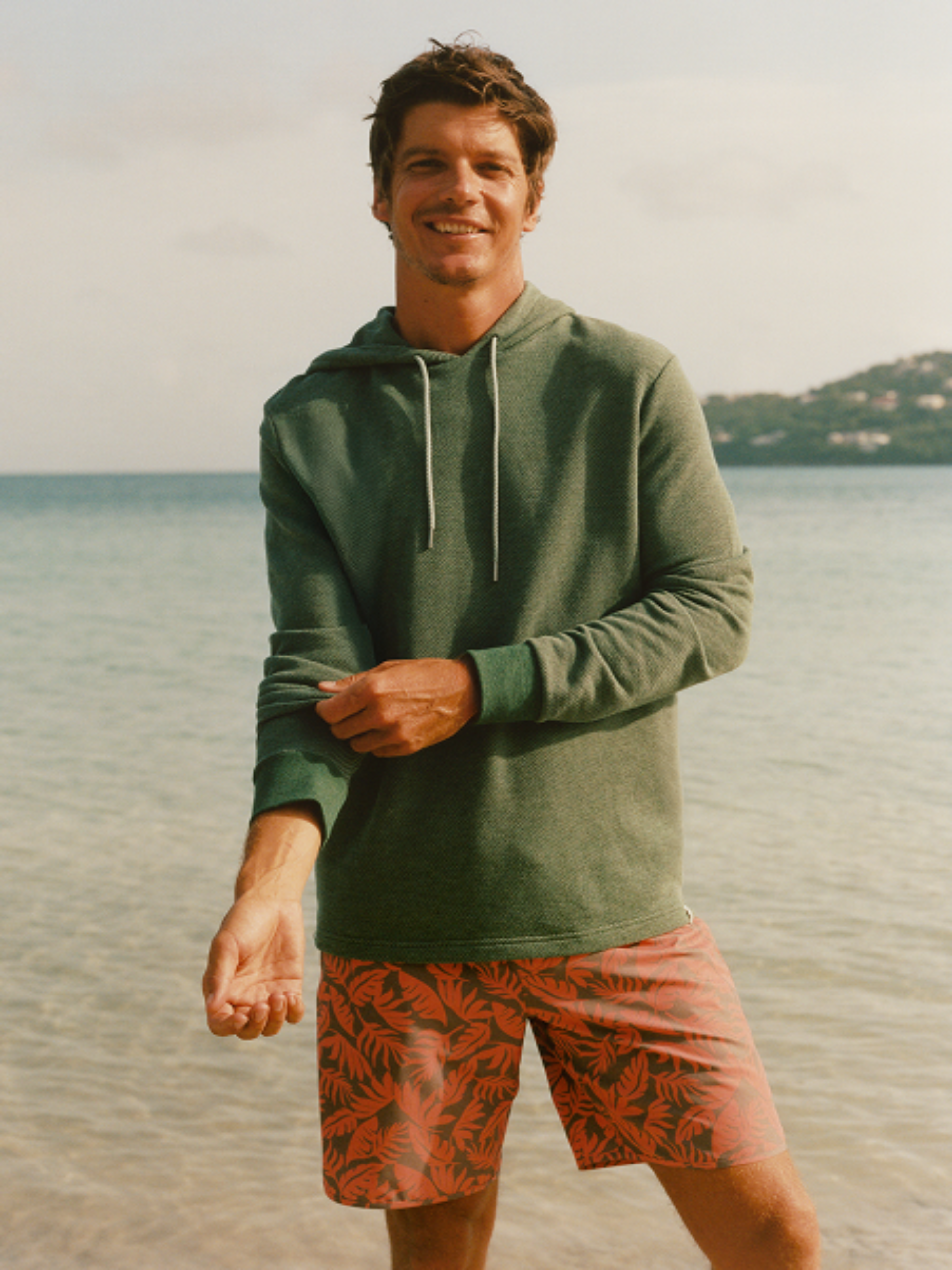 Man wearing a green tailwind hoodie and patterned anchor trunk standing by the water.