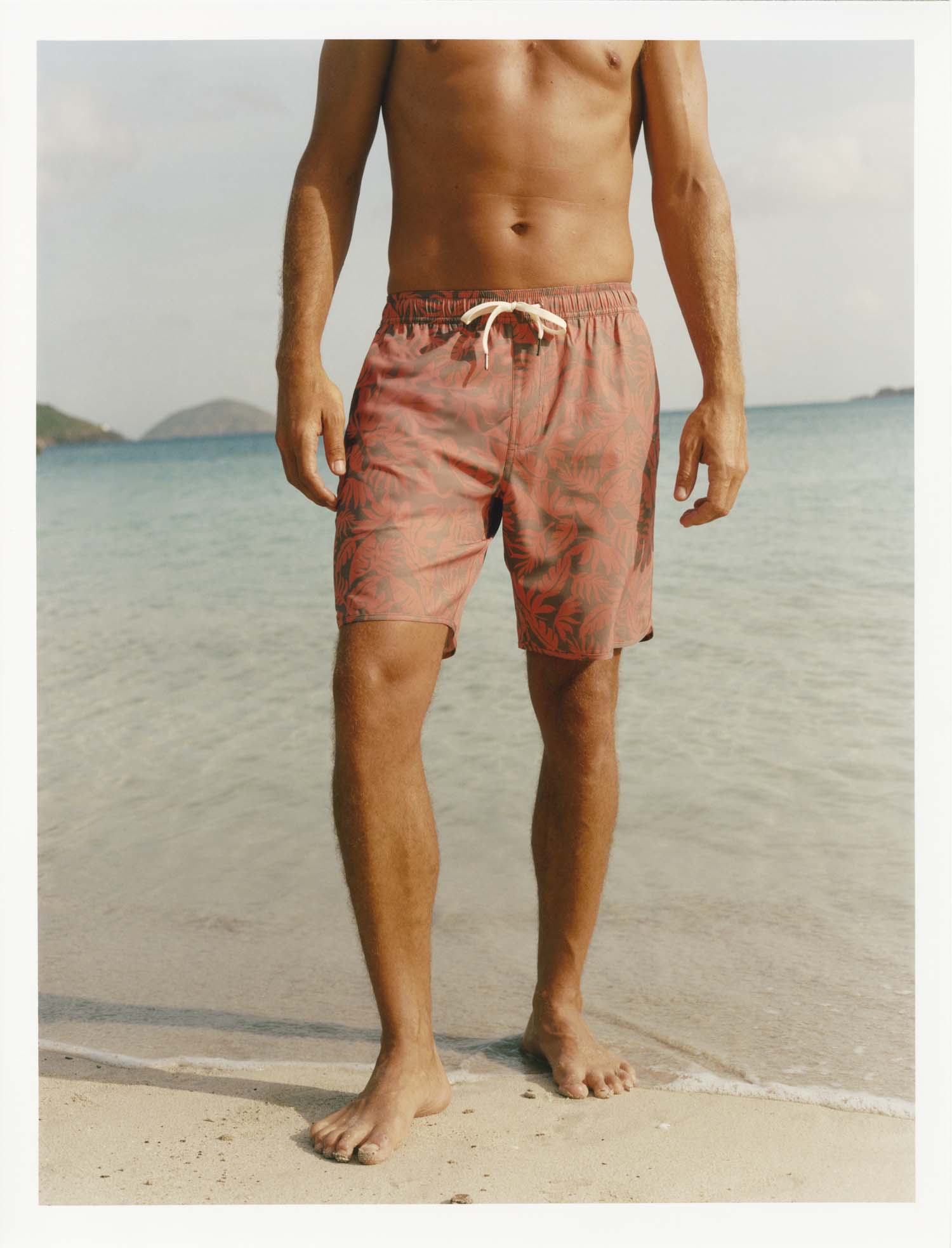 Person wearing orange and brown tropical leaves Anchor Trunk standing at edge of water on beach