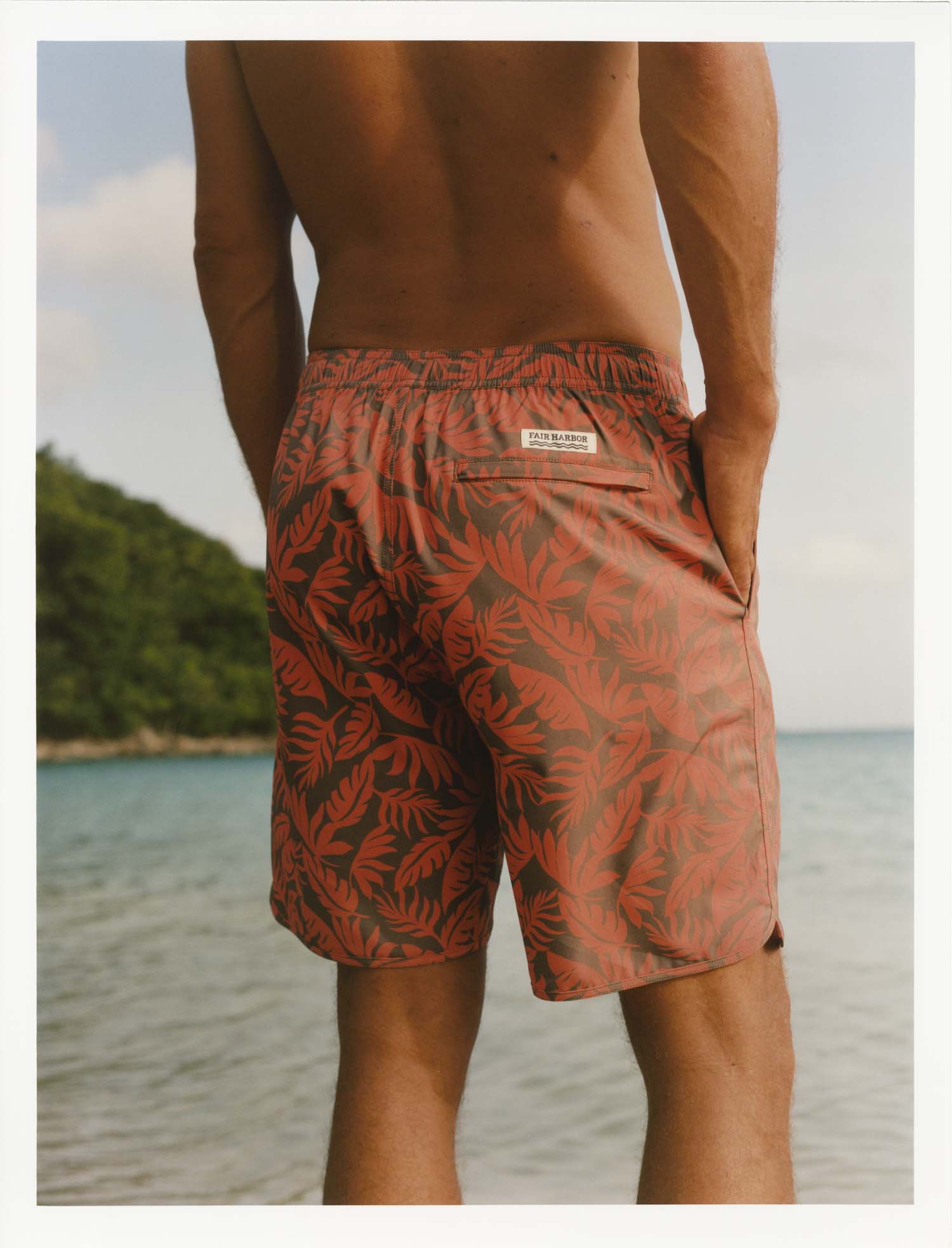 Person wearing orange and brown tropical leaves Anchor Trunk standing by a body of water with greenery in the background.