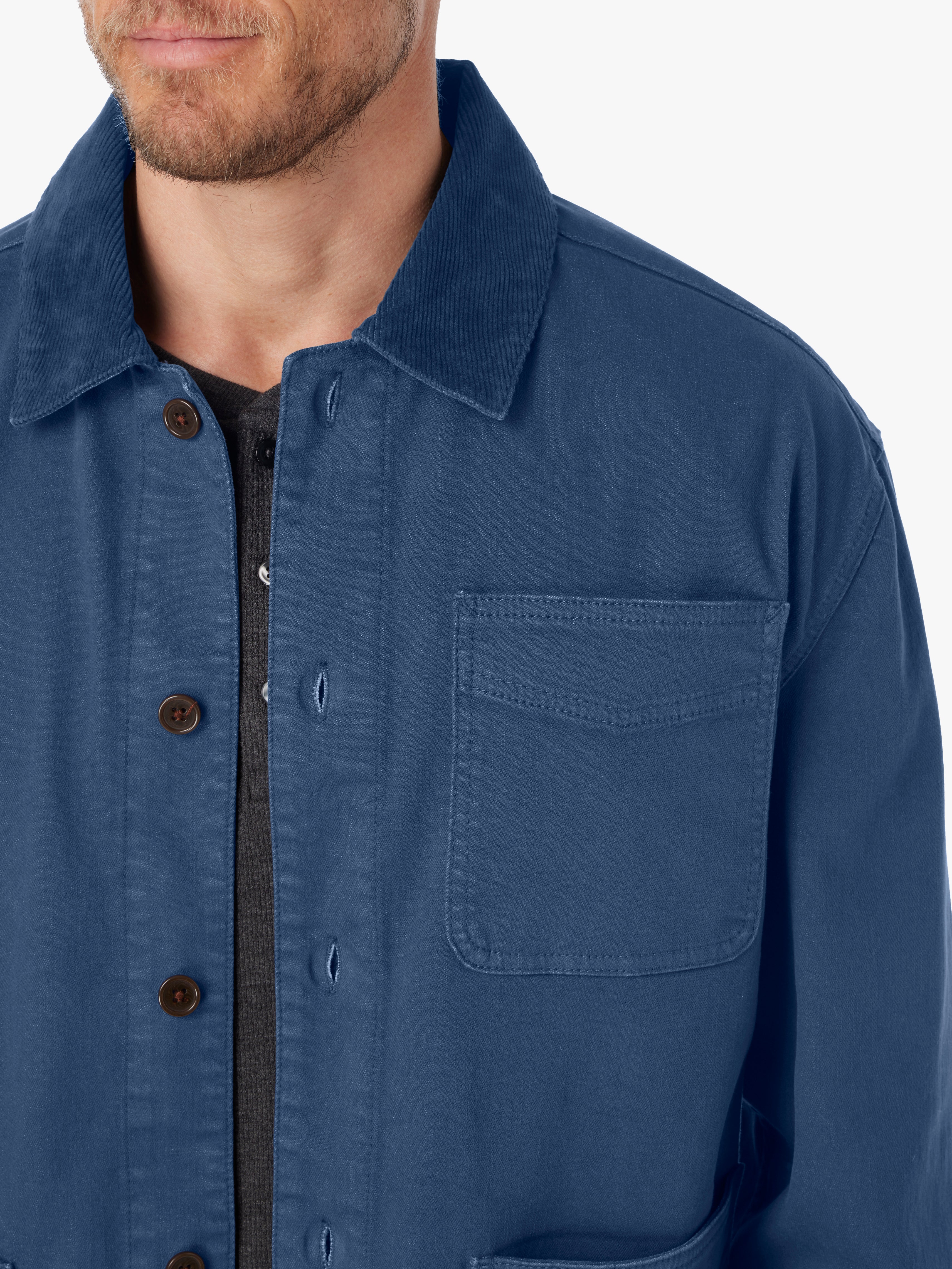 Close-up of man wearing blue barn style jacket with brown button details and tonal corduroy collar on a white background. 