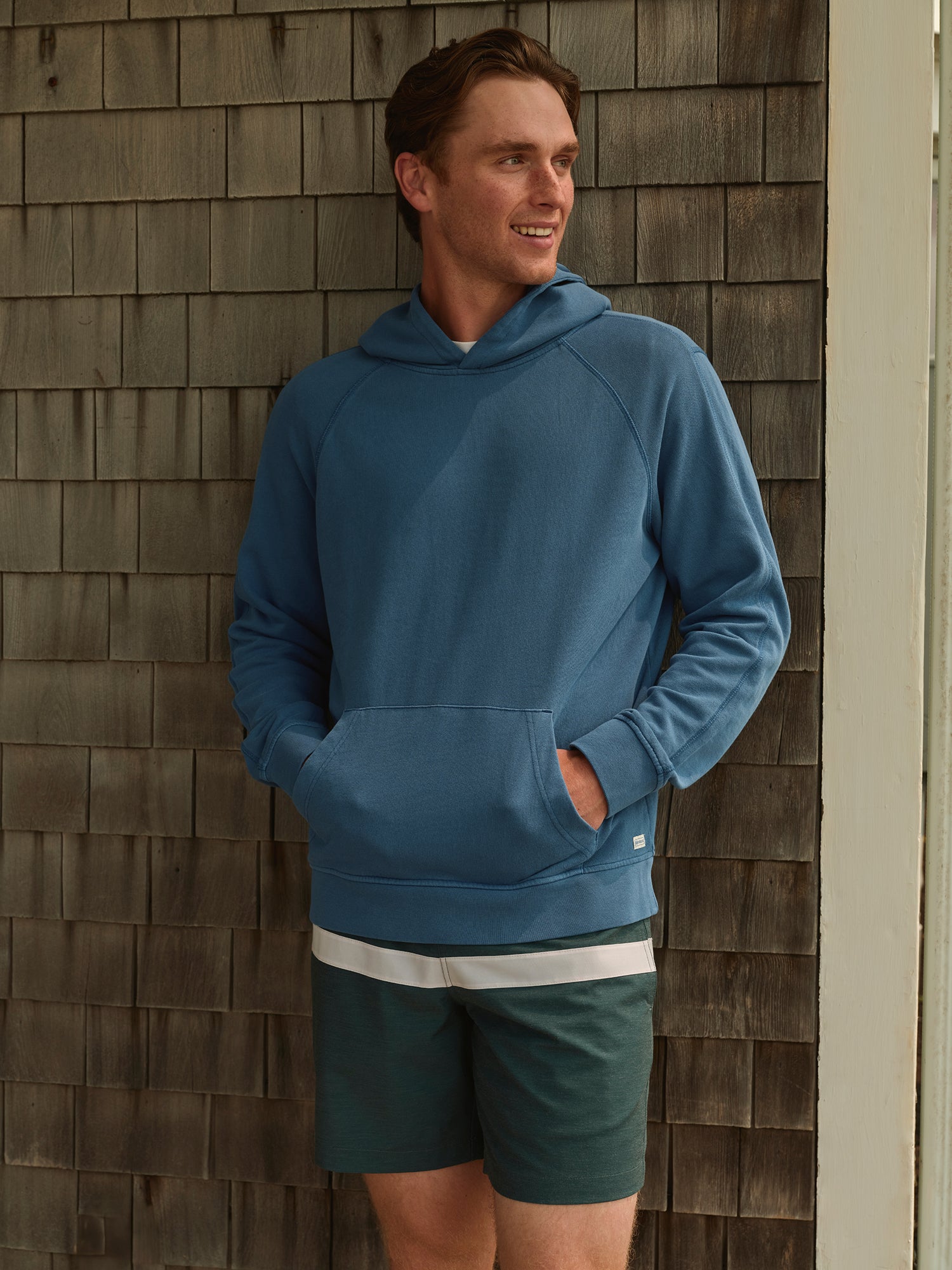 Man leaning on outside wall wearing blue hoodie with hands inside kangaroo pocket and green striped shorts