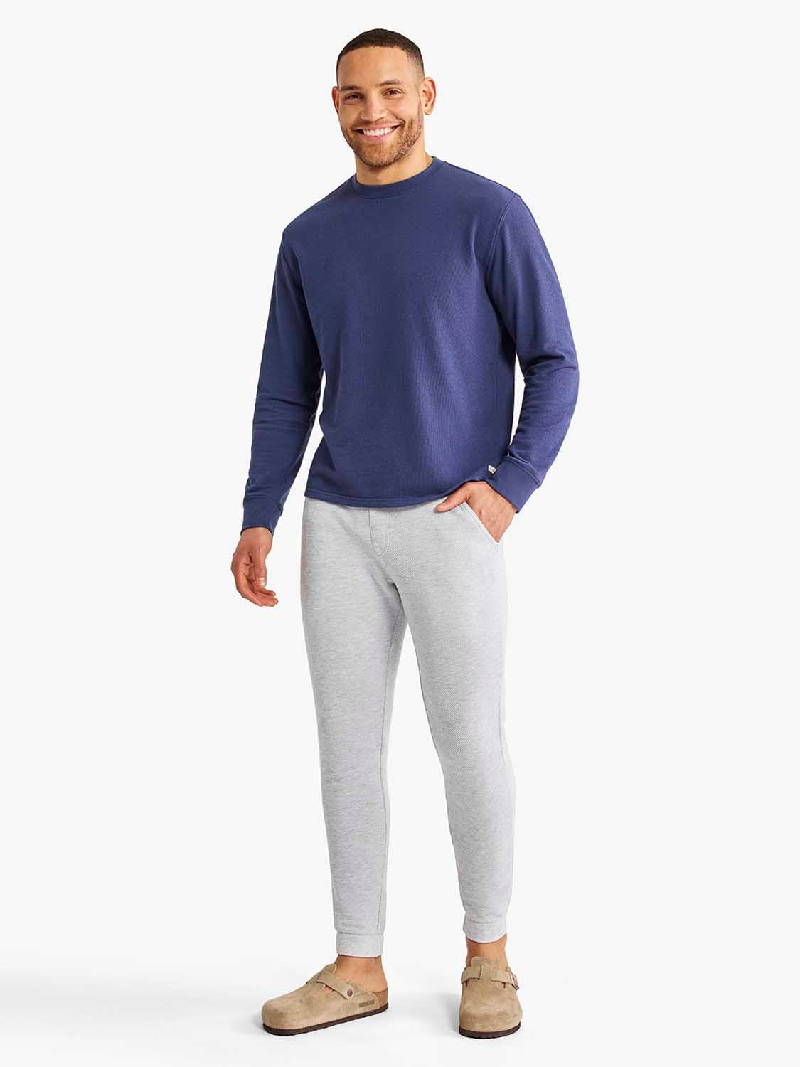 A man modeling a navy blue long-sleeve crewneck sweater and light gray heathered jogger pants with tan clogs, shot against a solid off-white studio background.