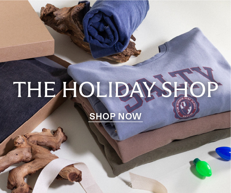 Stack of folded clothes, gift boxes and Christmas lights on a white surface, promotional text for 'The Holiday Shop Shop Now