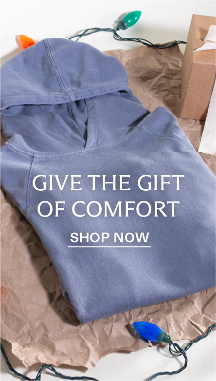Blue hoodie on brown wrapping paper with Christmas lights, text 'Give the Gift of Comfort Shop Now' on a white background.
